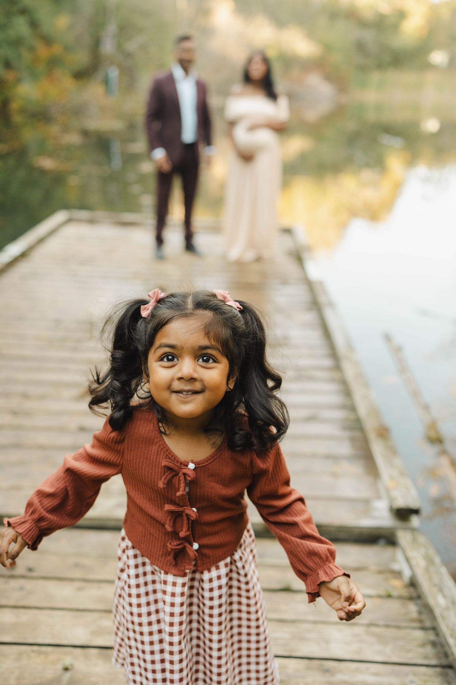Smiling little girl with pigtails runs along a wooden lakeside dock while her parents pose in the background during a maternity shoot.