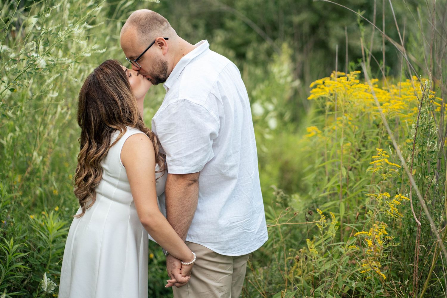 Couple in white shirt and white dress sharing a kiss while holding hands among yellow goldenrod wildflowers in a green outdoor field.