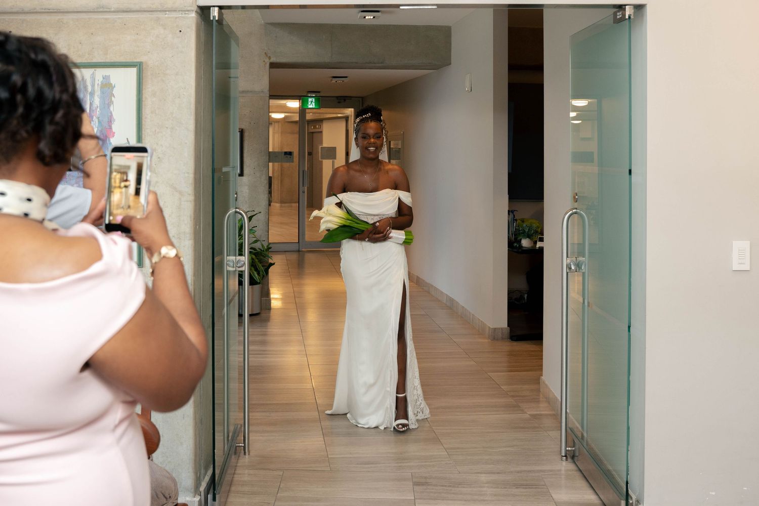 Bride in an off-shoulder white gown carries a bouquet of white flowers as she walks down a bright hallway while a guest takes a photo.