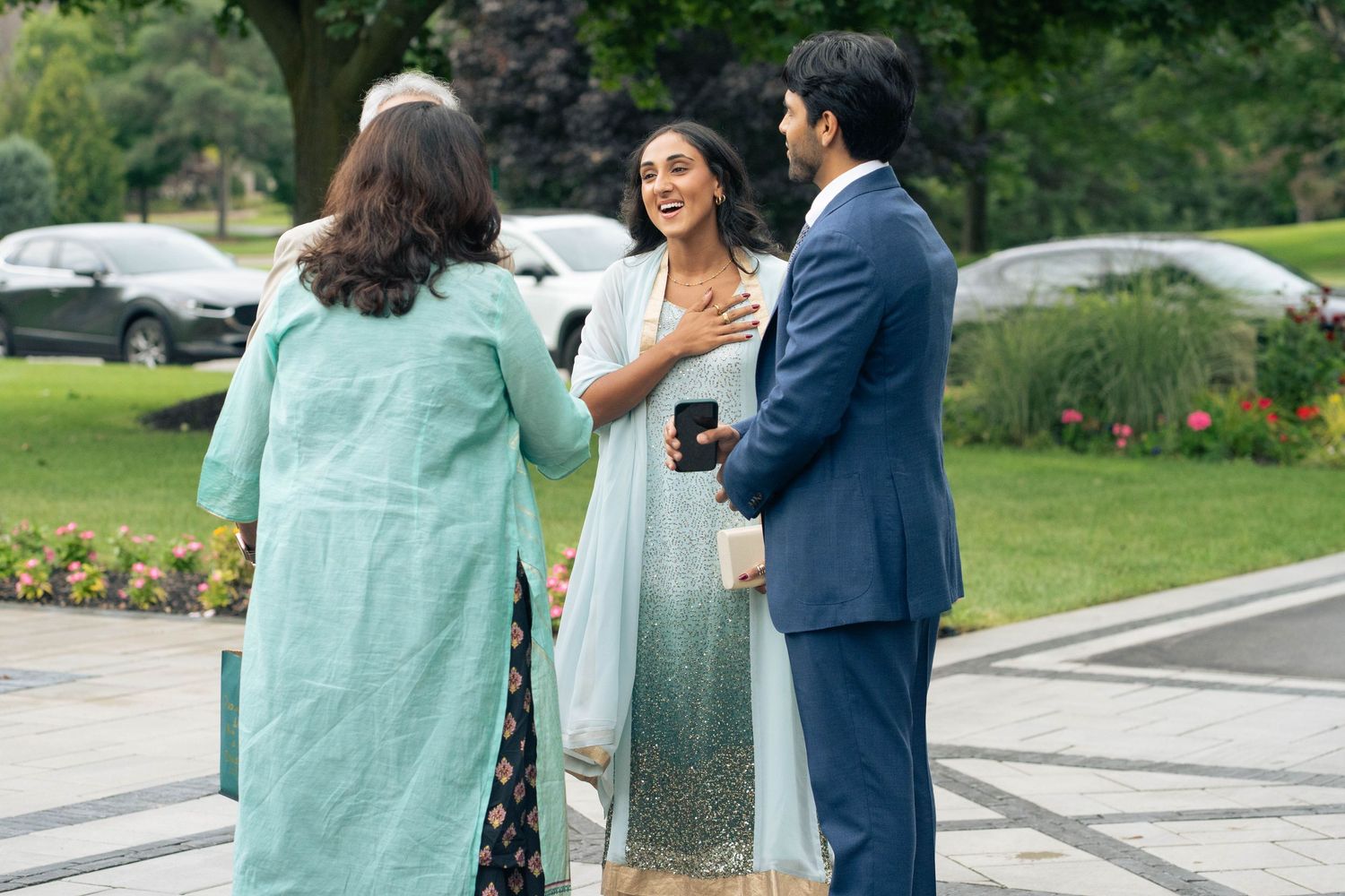Woman in a green gown places her hand on her chest while chatting with a man in a navy suit and another woman outdoors.