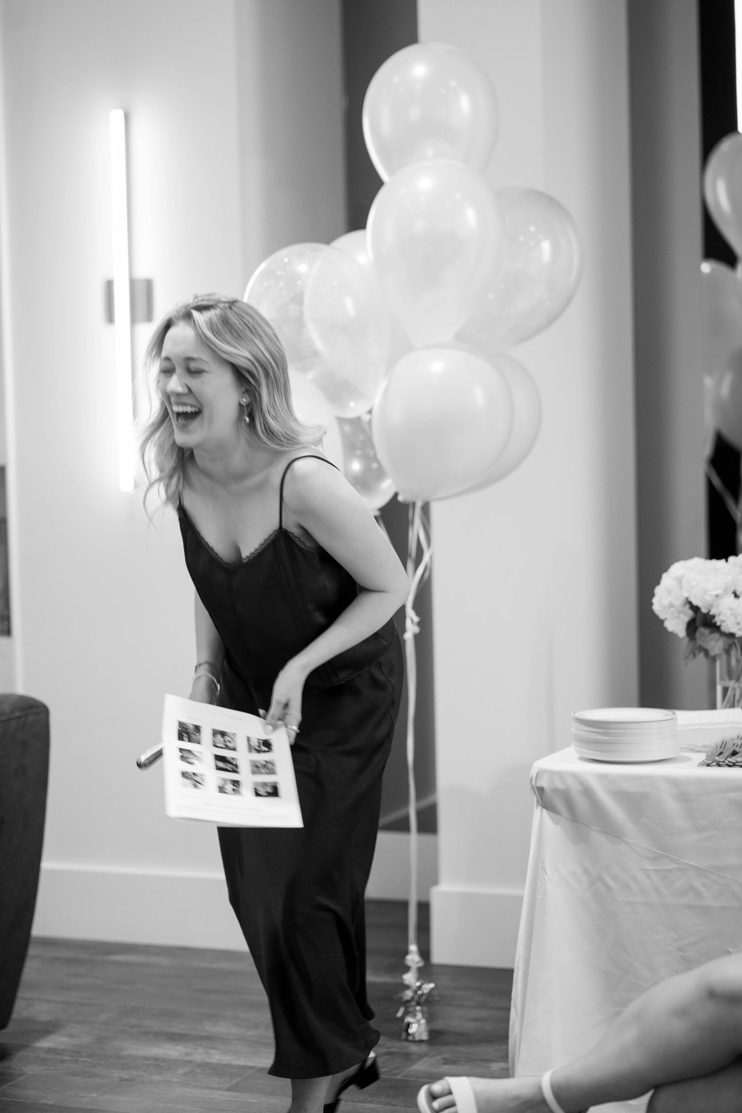 A woman in a satin slip dress laughs while holding a photo game sheet beside white balloons at an engagement party, in black and white.