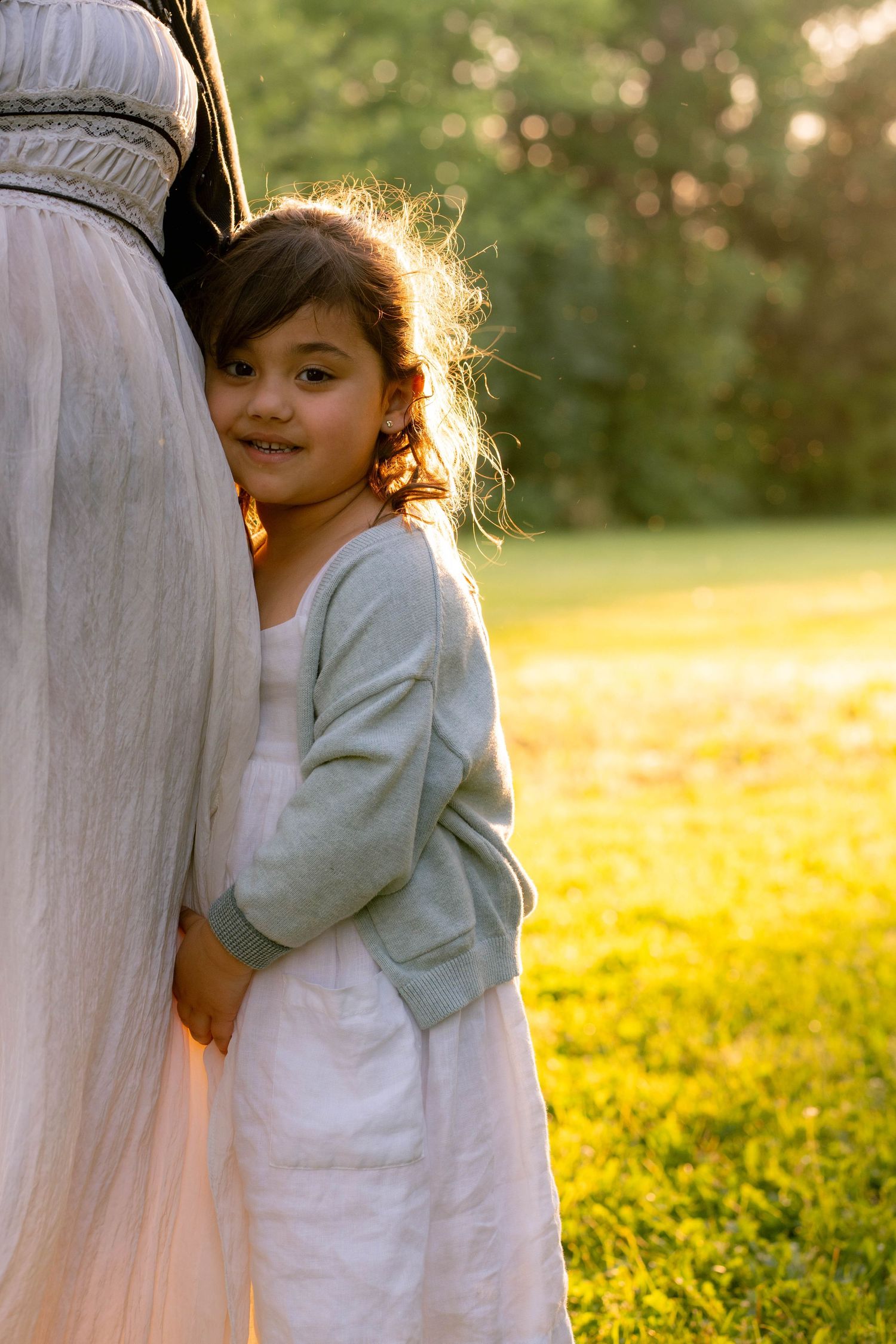 A smiling young girl in a sage green cardigan leans against her mother's white dress on a sunny green lawn.