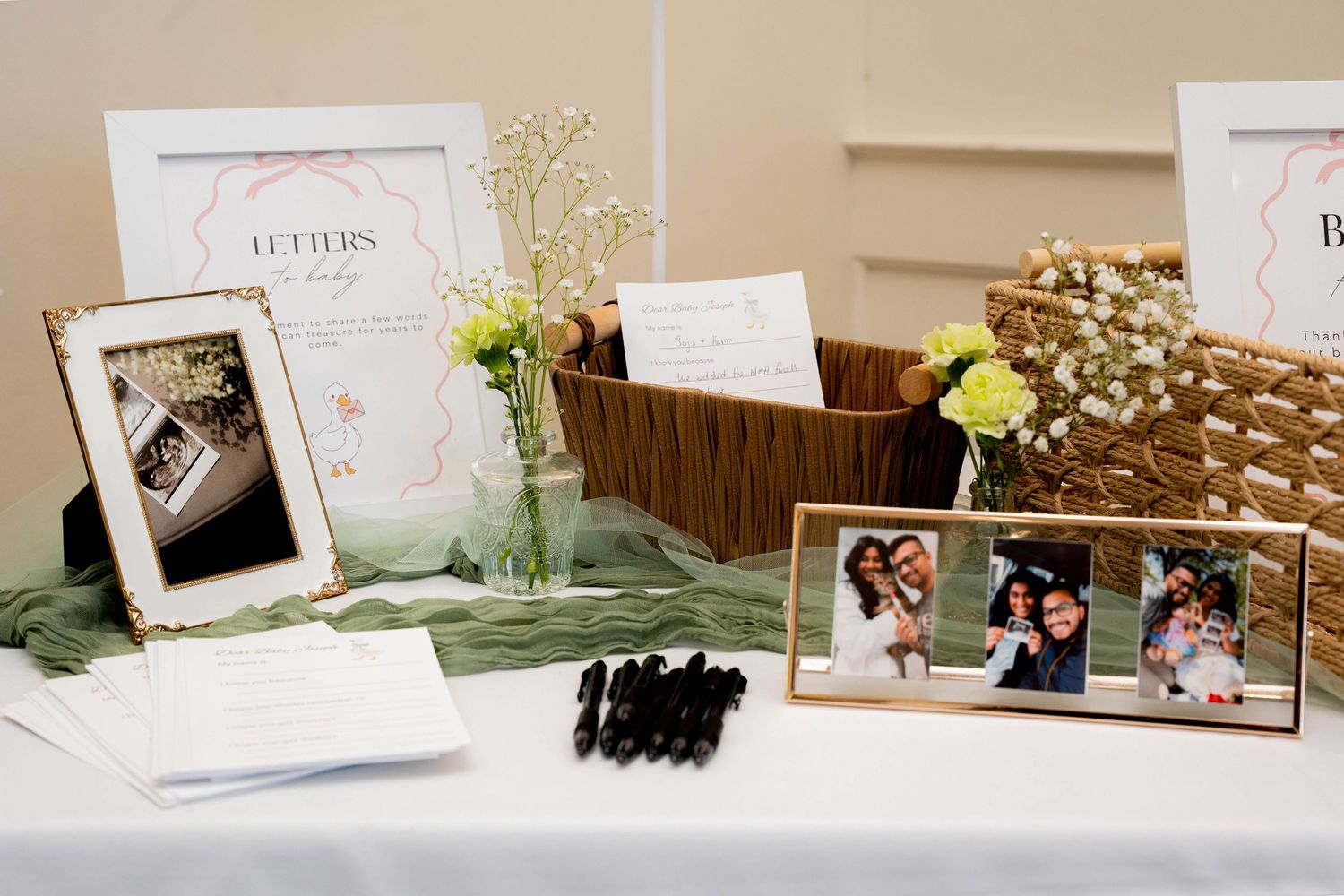 A baby shower guest table displays framed photos, a letters keepsake sign, yellow flowers, markers, and a woven basket on green fabric.