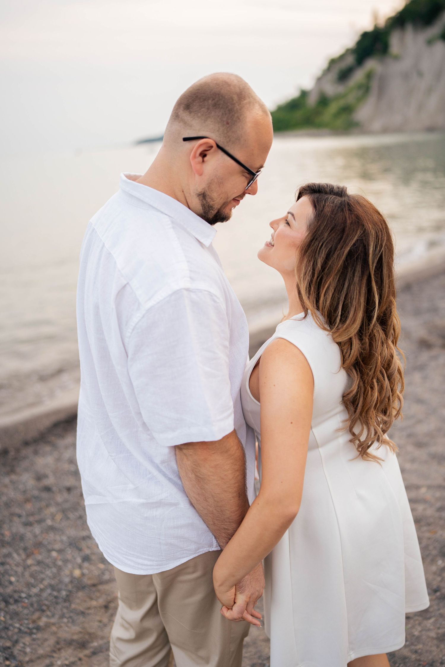 Couple in white outfits holding hands and smiling at each other on a pebble beach near scenic bluffs at sunset.
