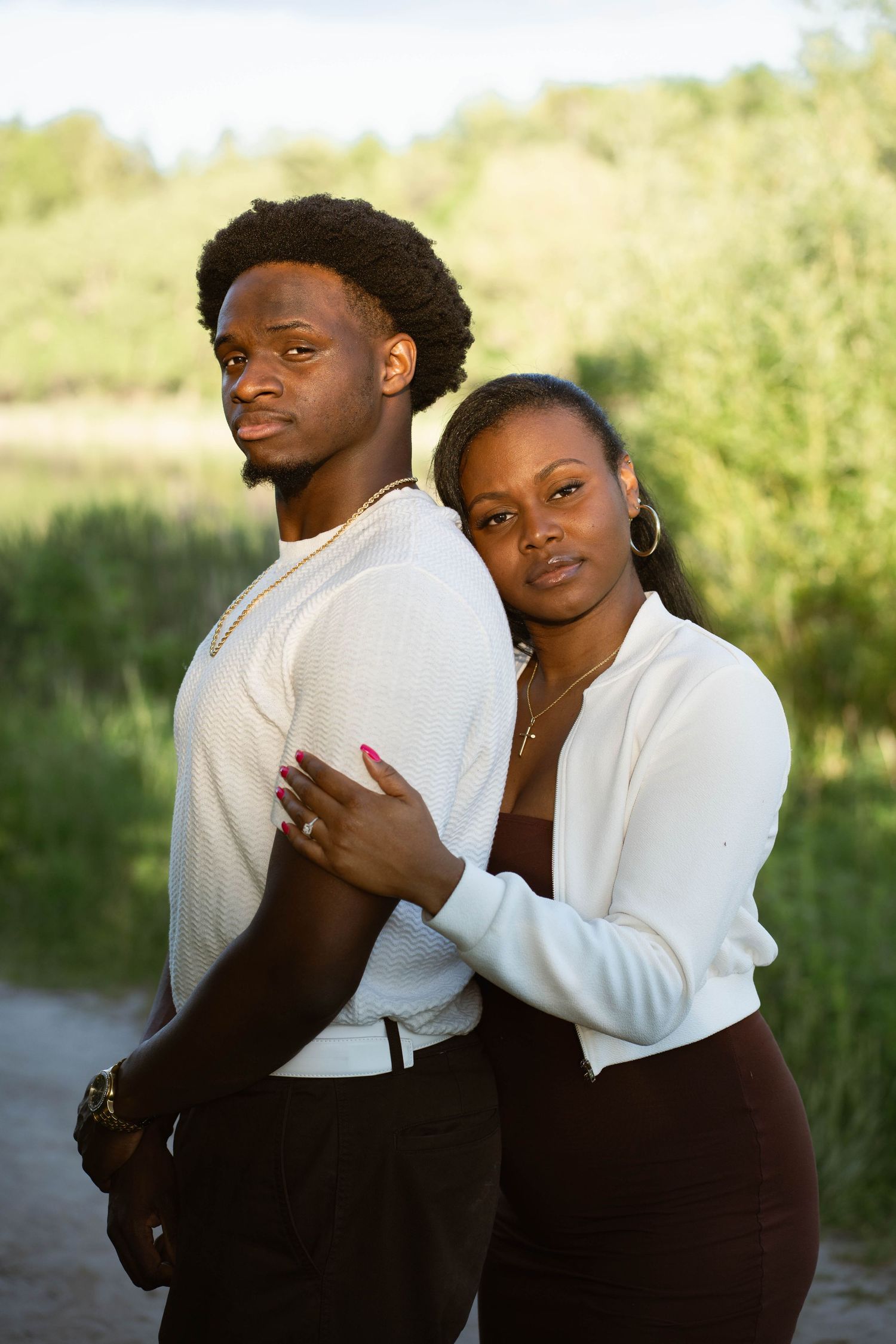 Romantic engagement portrait of a couple embracing outdoors, the woman showing her ring beside a grassy lakeside.