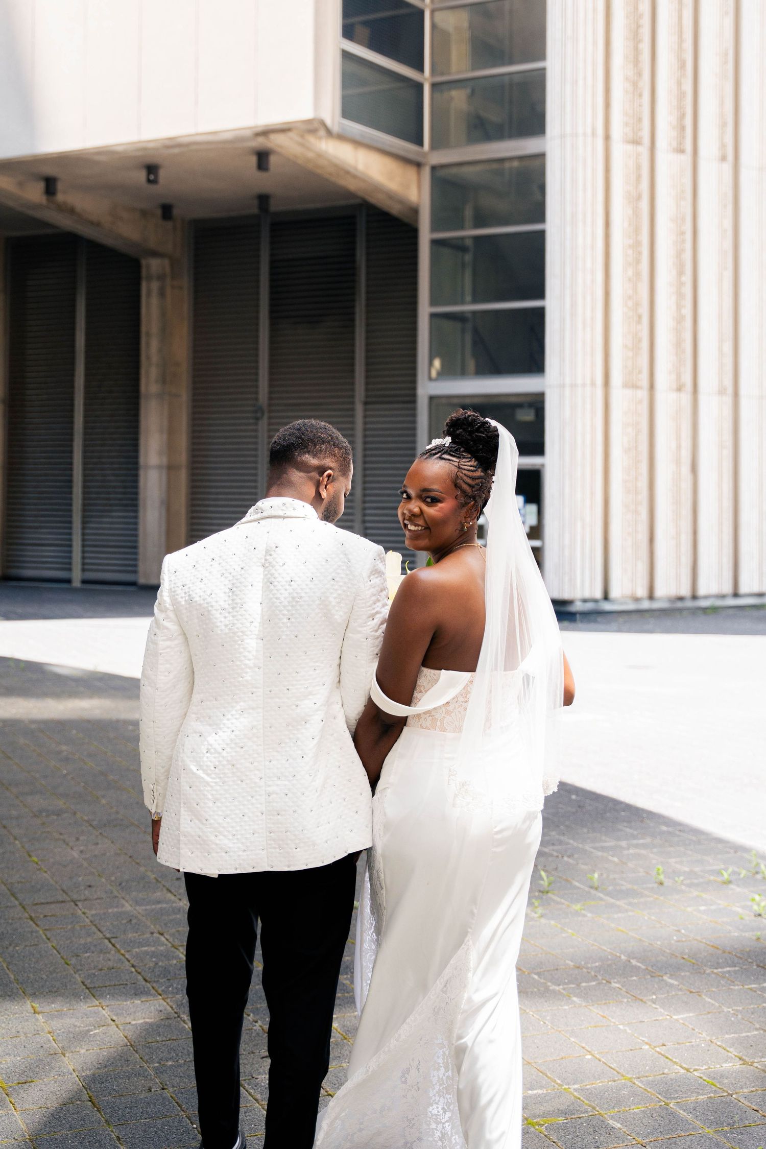 Newlyweds walk hand in hand toward a modern building, the bride in a long veil and white gown, the groom in an ivory jacket.