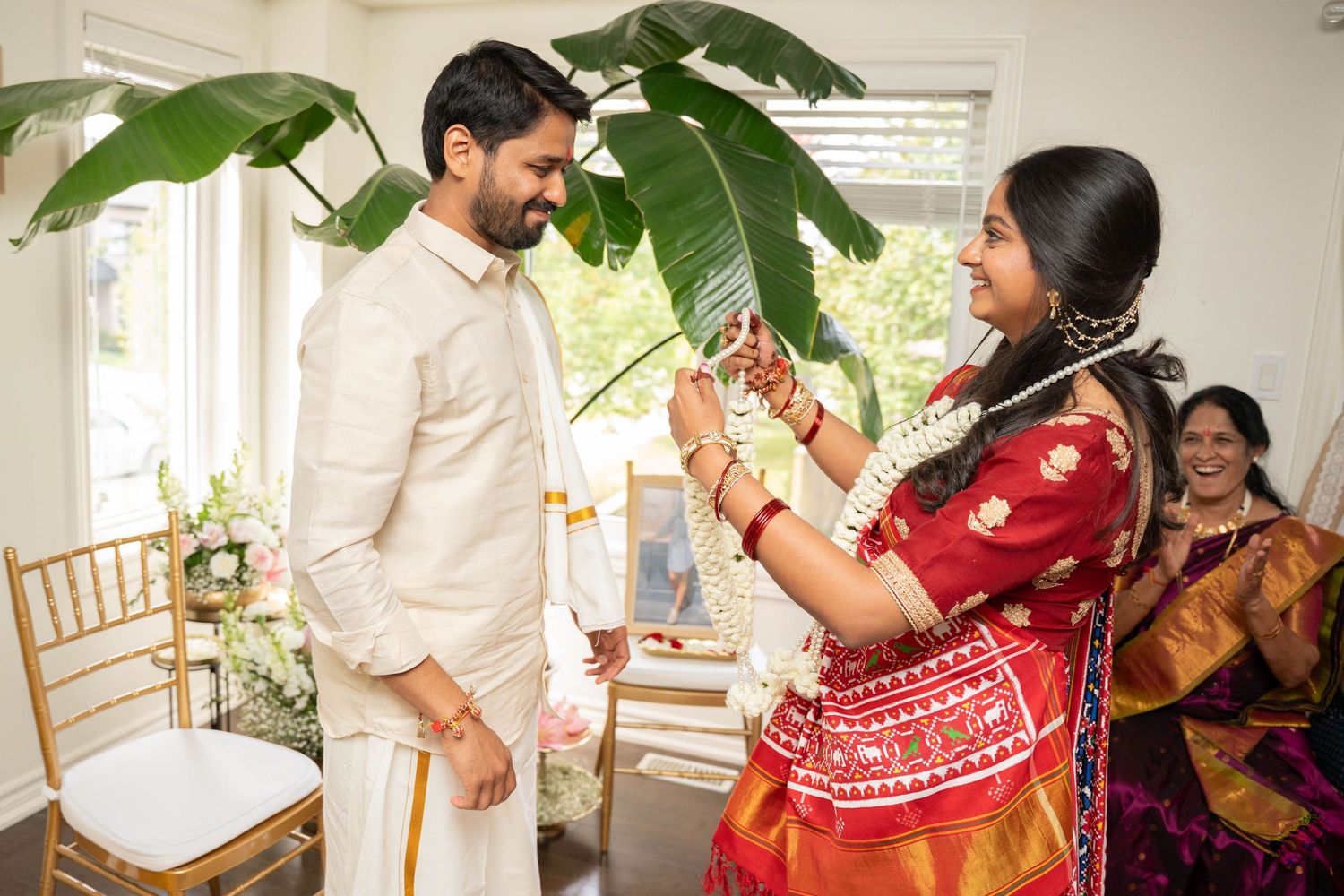 Bride in a red and orange silk saree exchanges flower garlands with a groom in a cream traditional outfit during an Indian wedding ceremony.