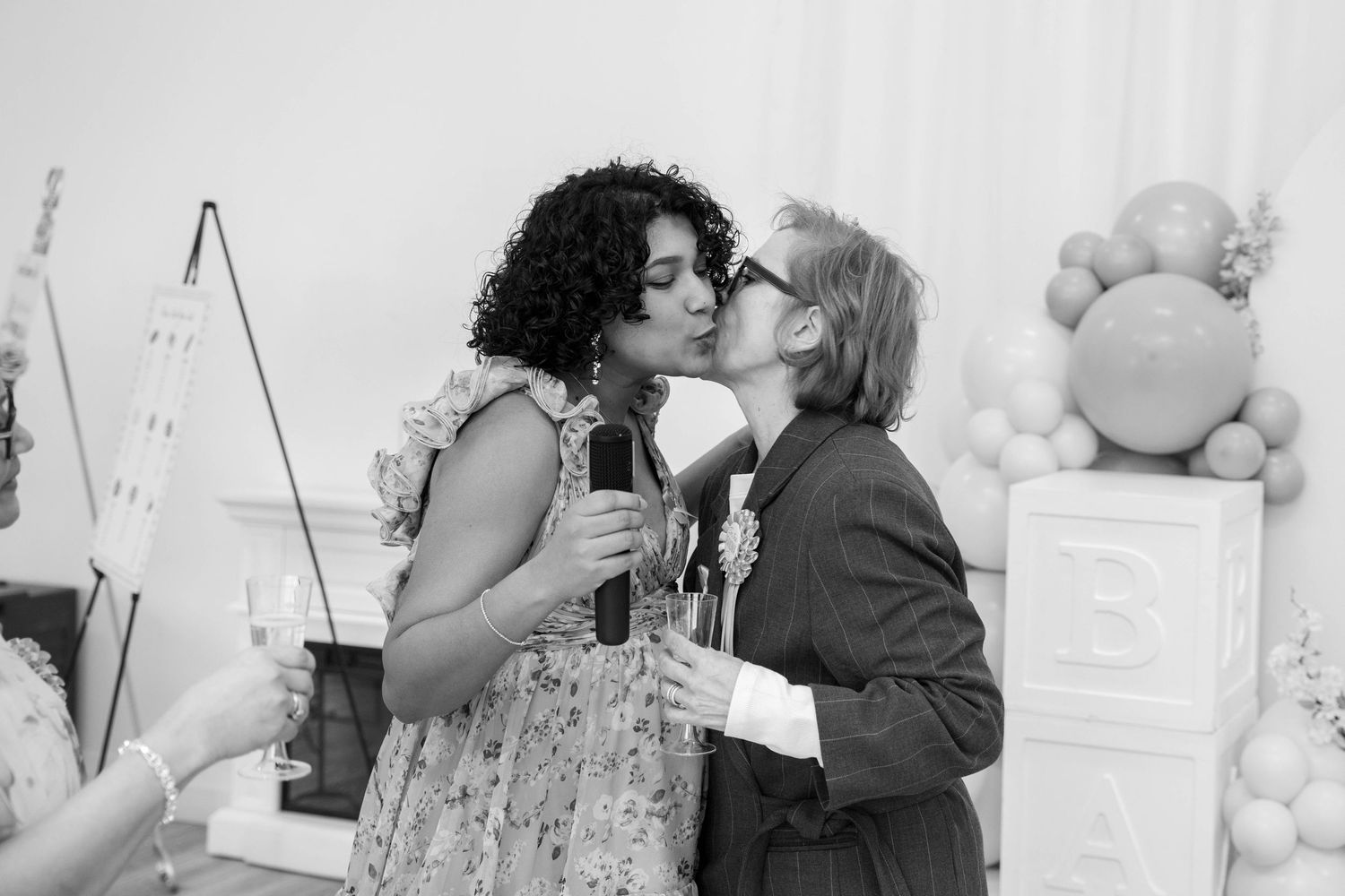 Black and white photo of a pregnant woman holding a microphone as a woman kisses her cheek at a baby shower.