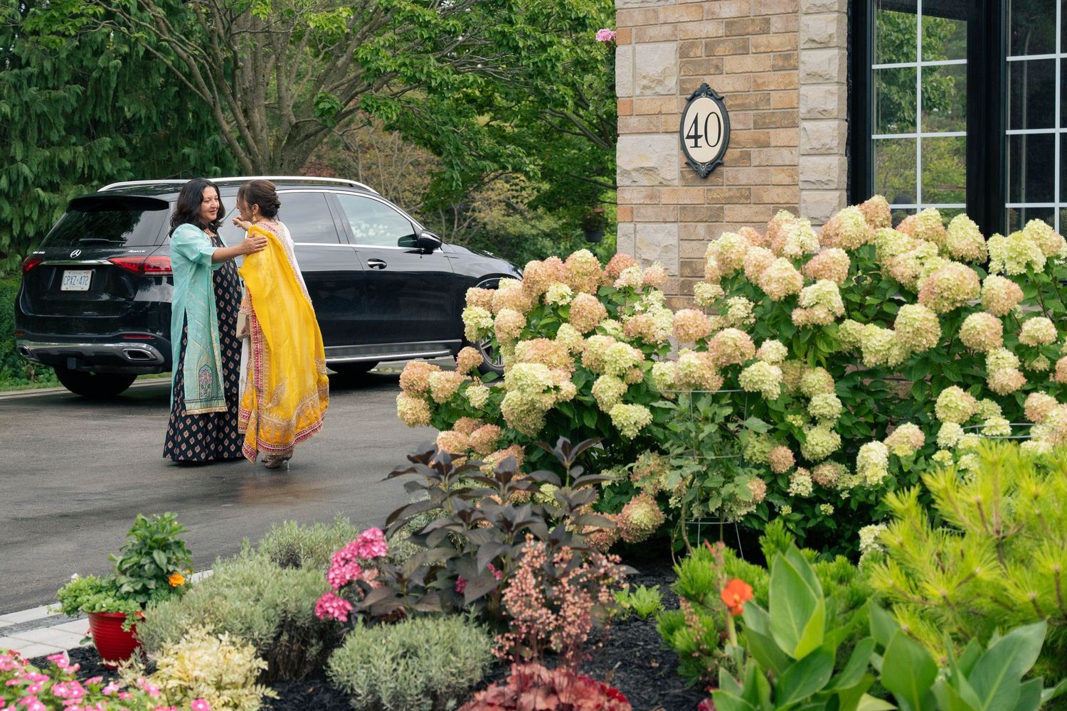 Two women in colorful traditional saris greet each other beside blooming white hydrangeas near a brick house entrance.