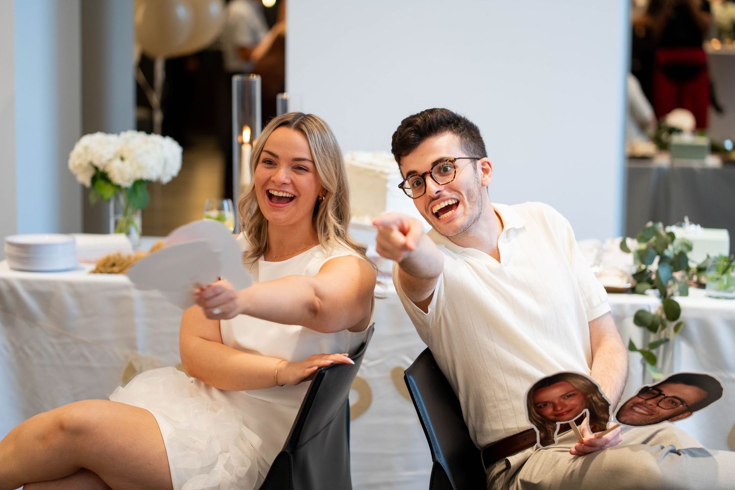 A laughing woman holds a game sheet while a man in glasses points playfully, with face cutouts on his lap at an engagement party.