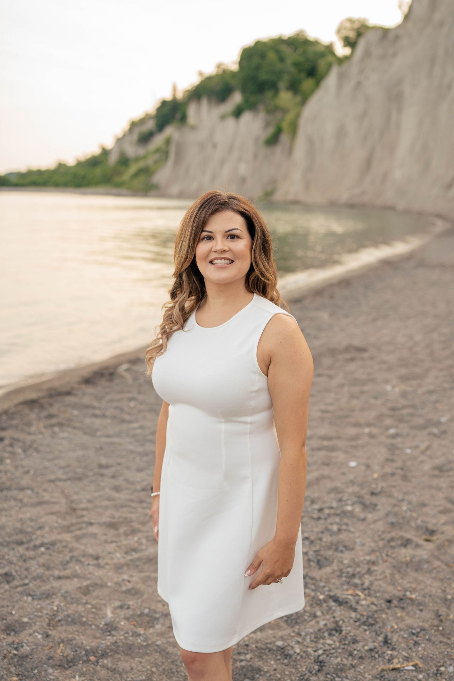 Smiling woman in a white sleeveless dress standing on a pebbly beach with tall sandy bluffs behind her at sunset.
