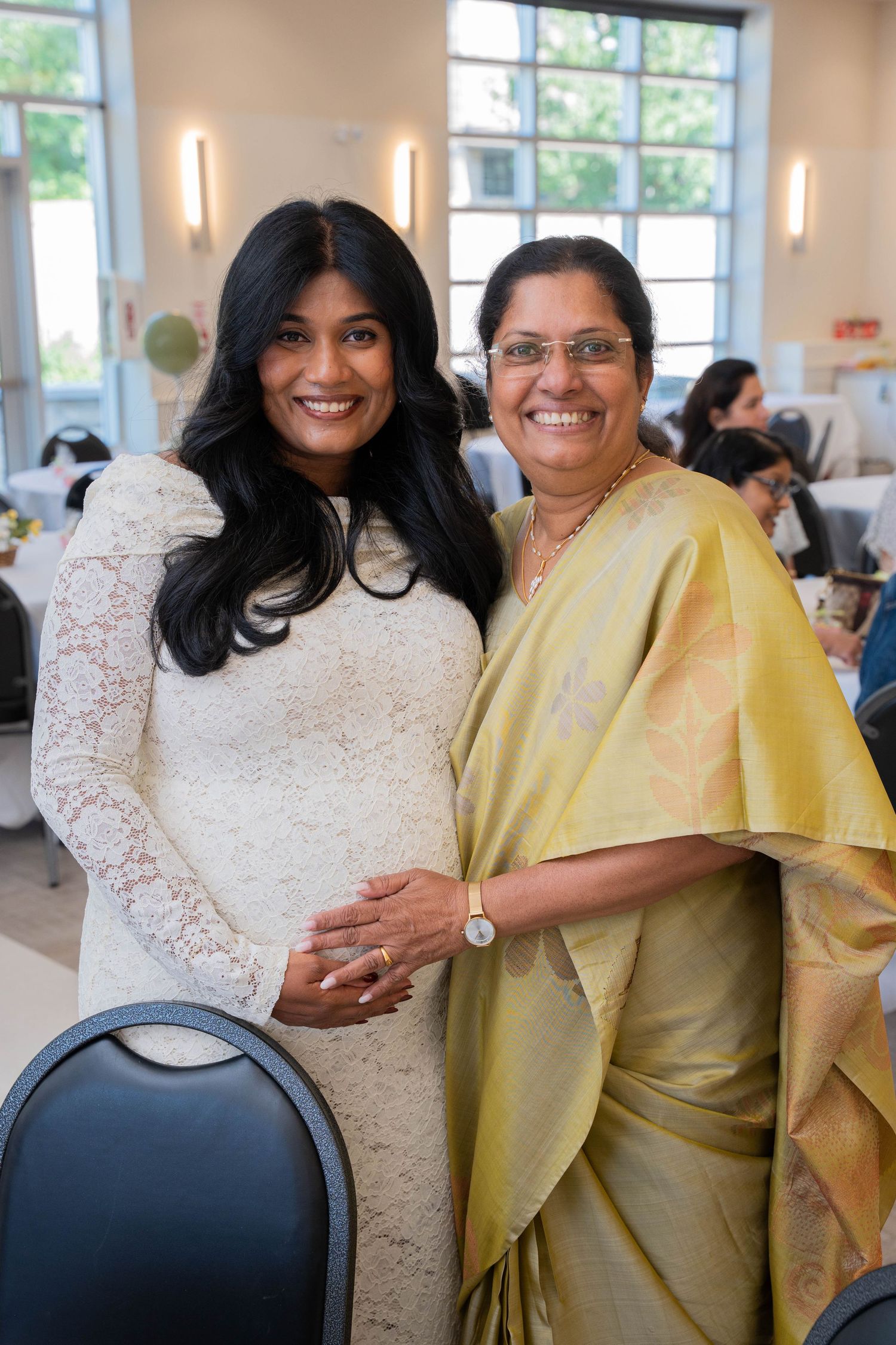 Expectant mother in a white lace dress poses with a smiling older woman in a golden sari, hands resting on her baby bump.