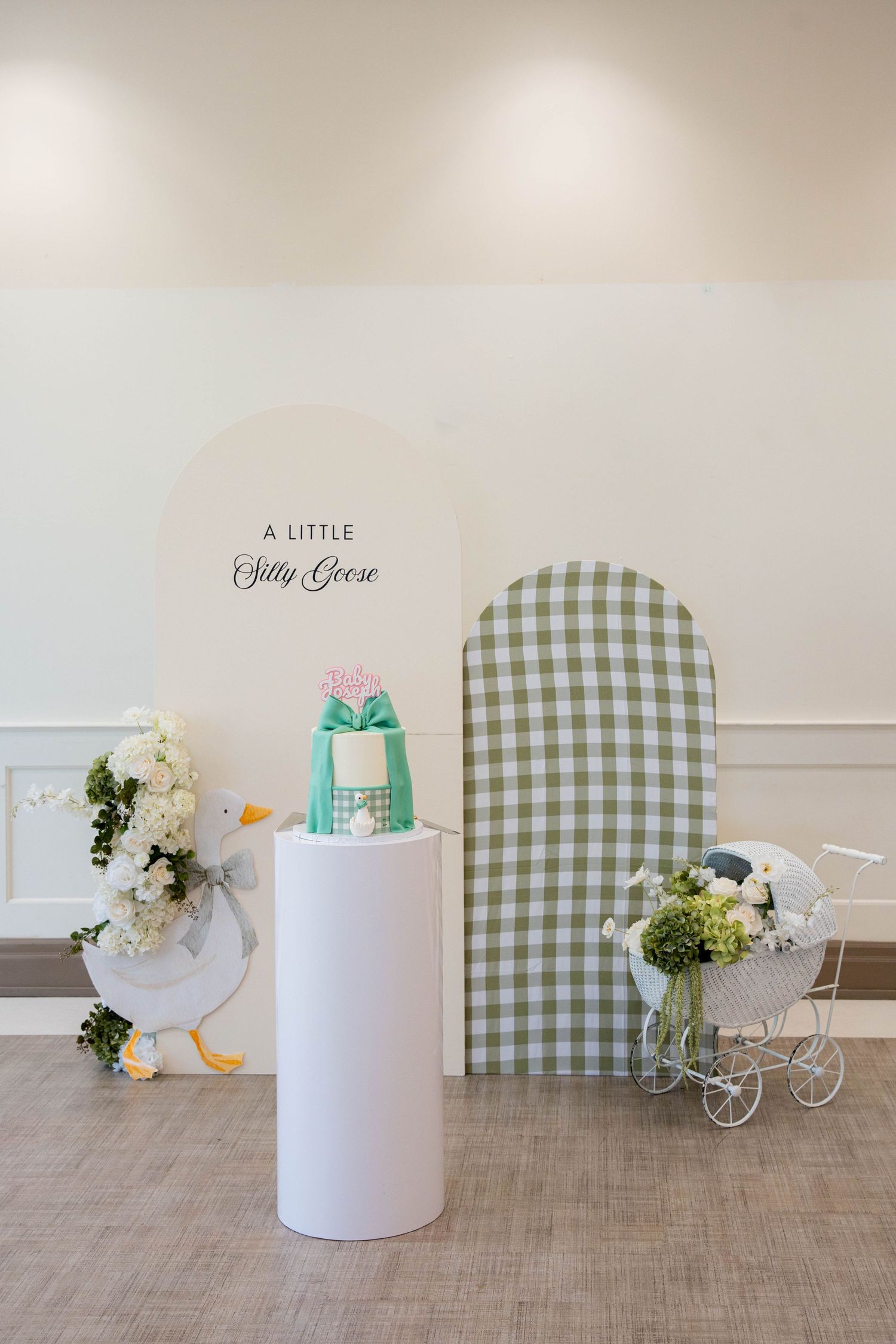 Baby shower display with a mint green bow cake on a white pedestal, a silly goose sign, floral goose cutout, gingham arch and white pram.