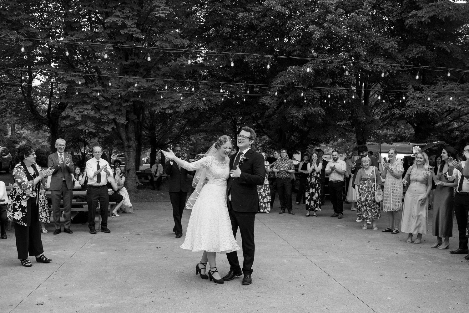 Black and white photo of the bride and groom sharing a first dance on a patio as guests watch beneath string lights and trees.