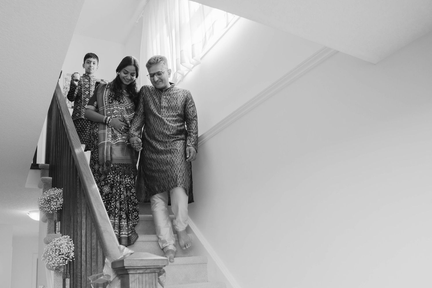 Black and white photo of a man in a patterned kurta and a woman walking down a staircase together, with another person behind them.