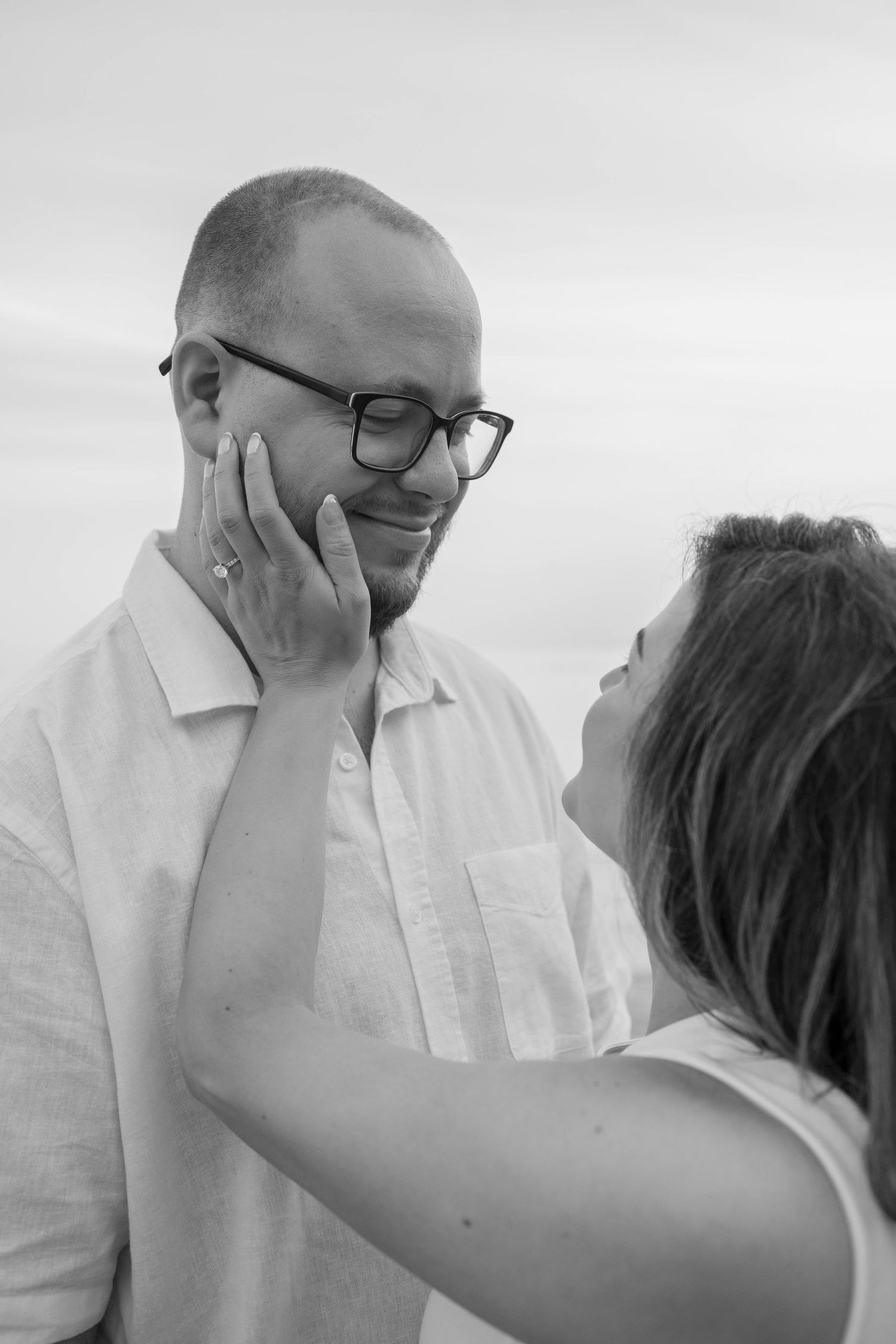 Black and white engagement photo of a woman gently touching a smiling bearded man's face, her ring visible.