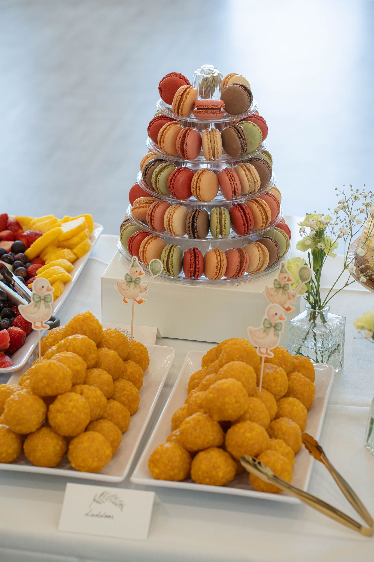 Dessert table with a tower of colorful macarons, golden treats, fresh fruit platters, and goose-themed decorative picks.