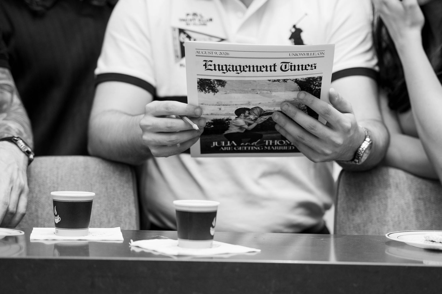 Black and white photo of guests holding an Engagement Times newspaper at a table with two coffee cups.