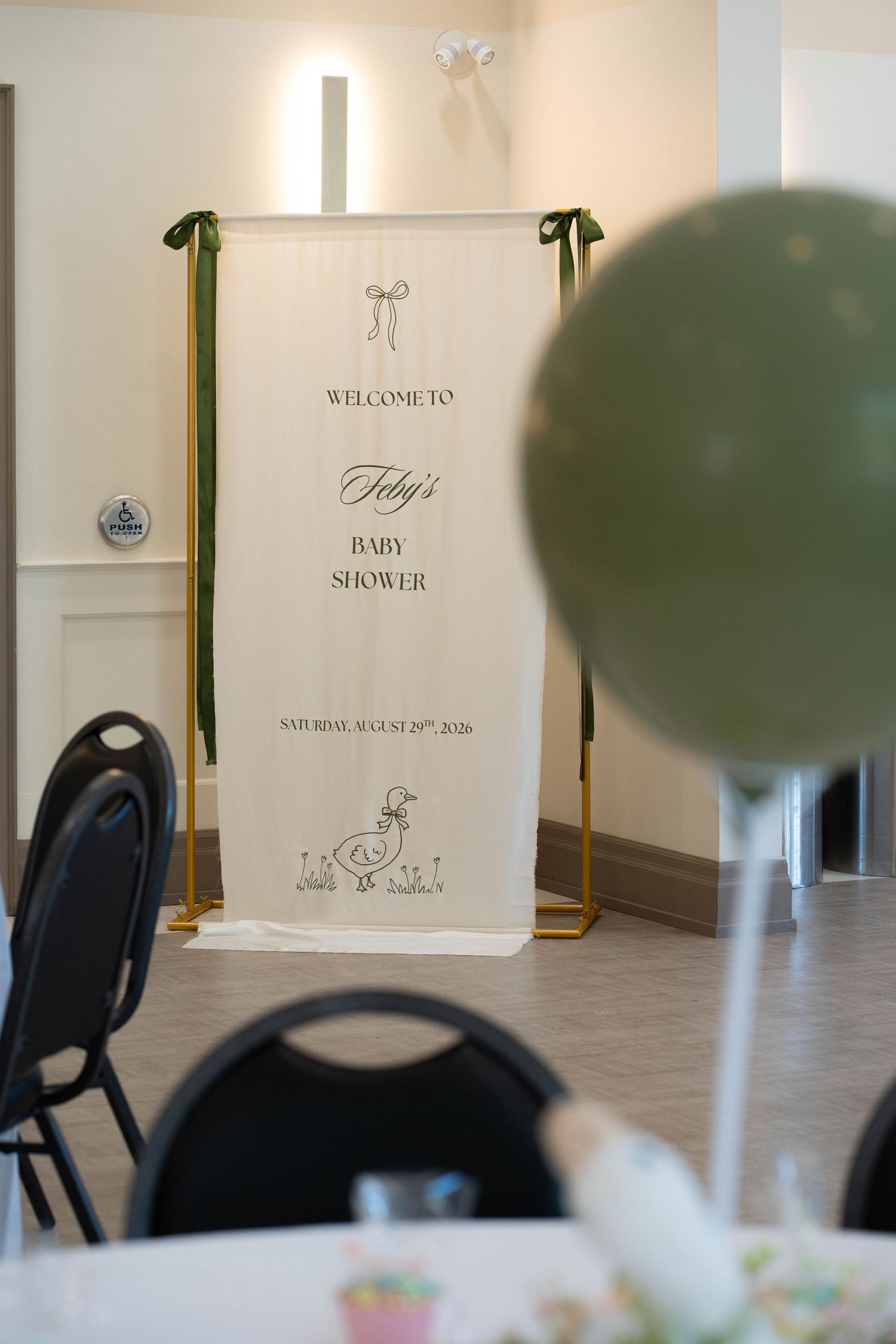 A welcome sign at a baby shower with a duck illustration stands behind a green balloon and black chairs in a bright event hall.