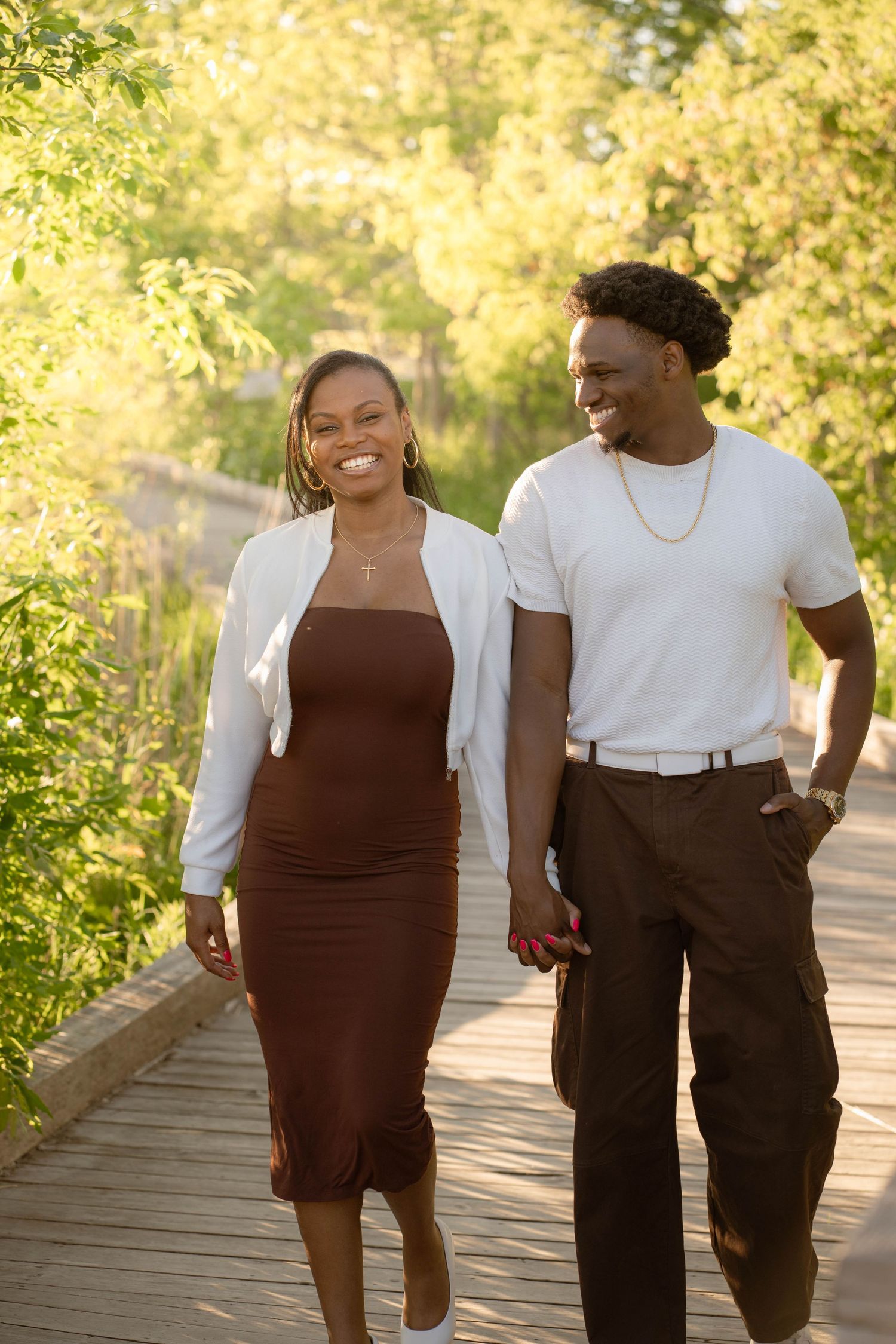 Happy couple laughing and holding hands while strolling across a wooden boardwalk lined with lush summer greenery.