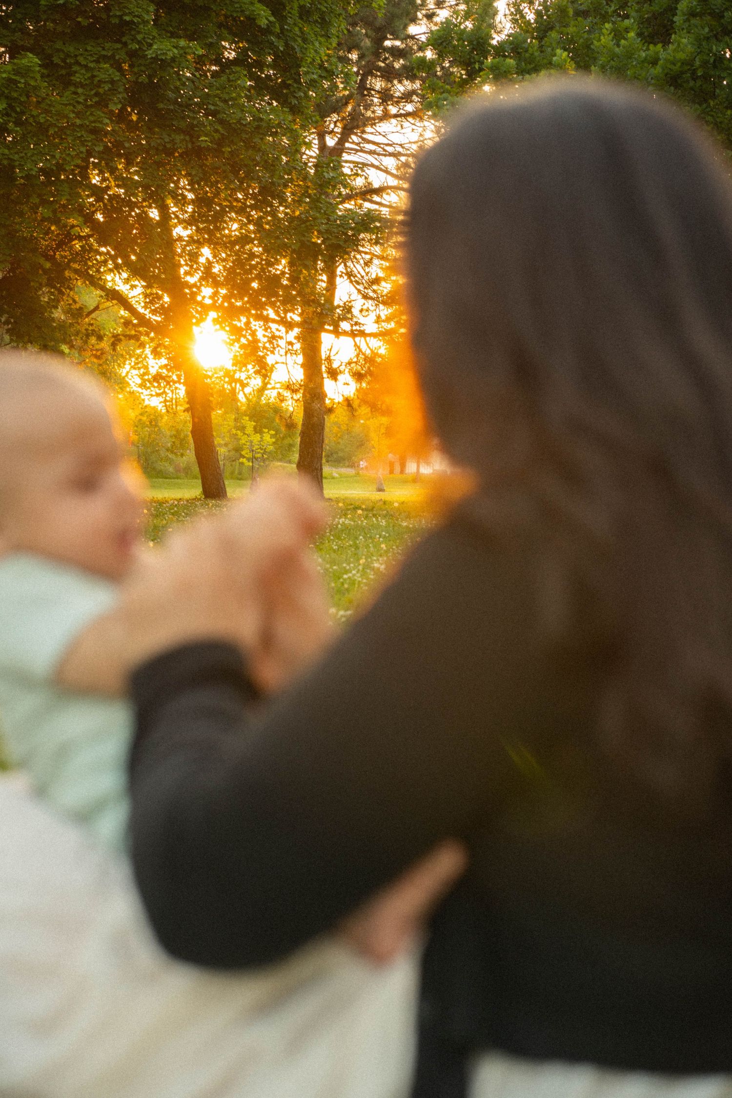 A blurred mother holds her baby in the foreground as golden sunset light glows through the trees in a park.