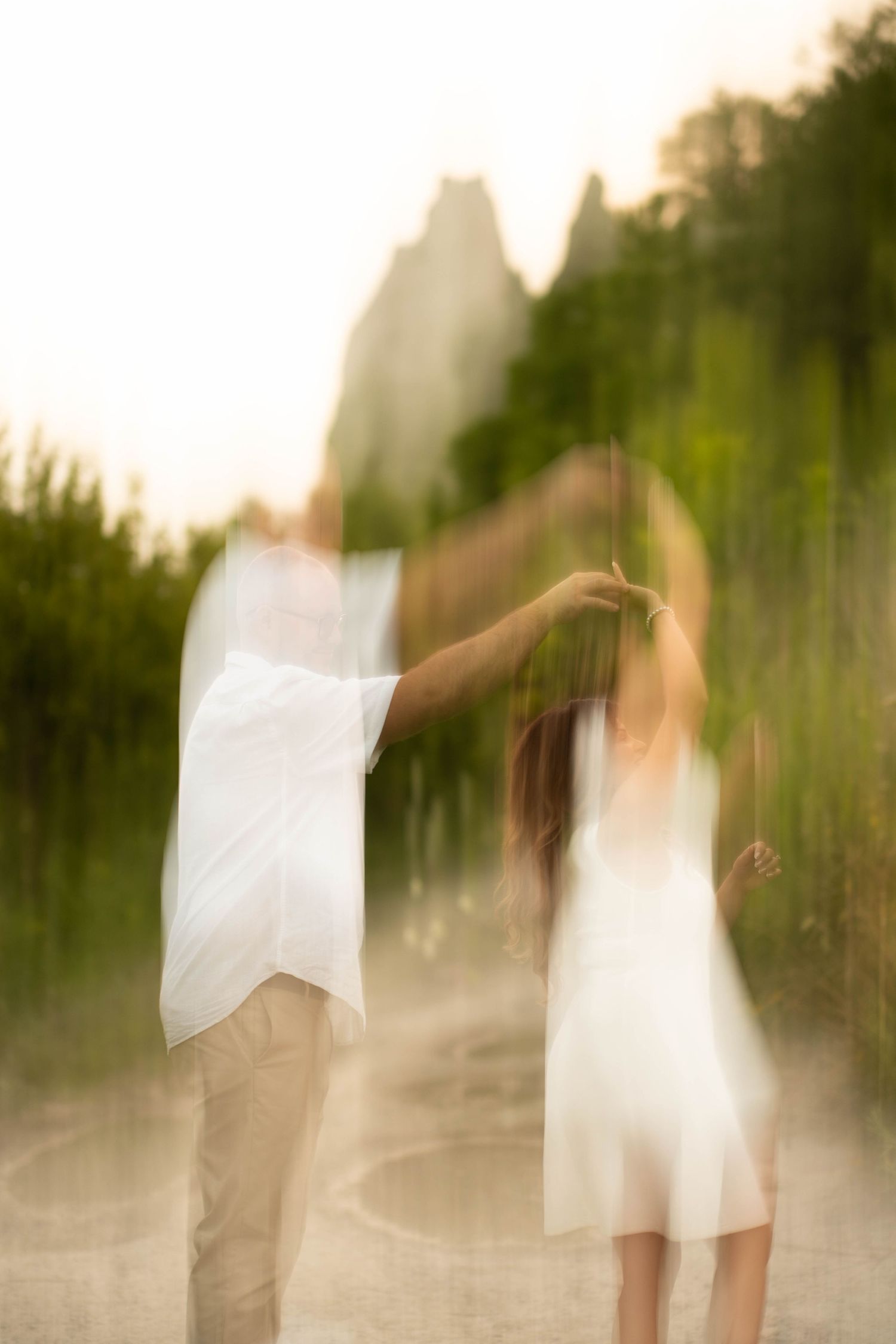 Artistic motion-blur photo of a couple dancing in white and khaki outfits with scenic bluffs in the background.