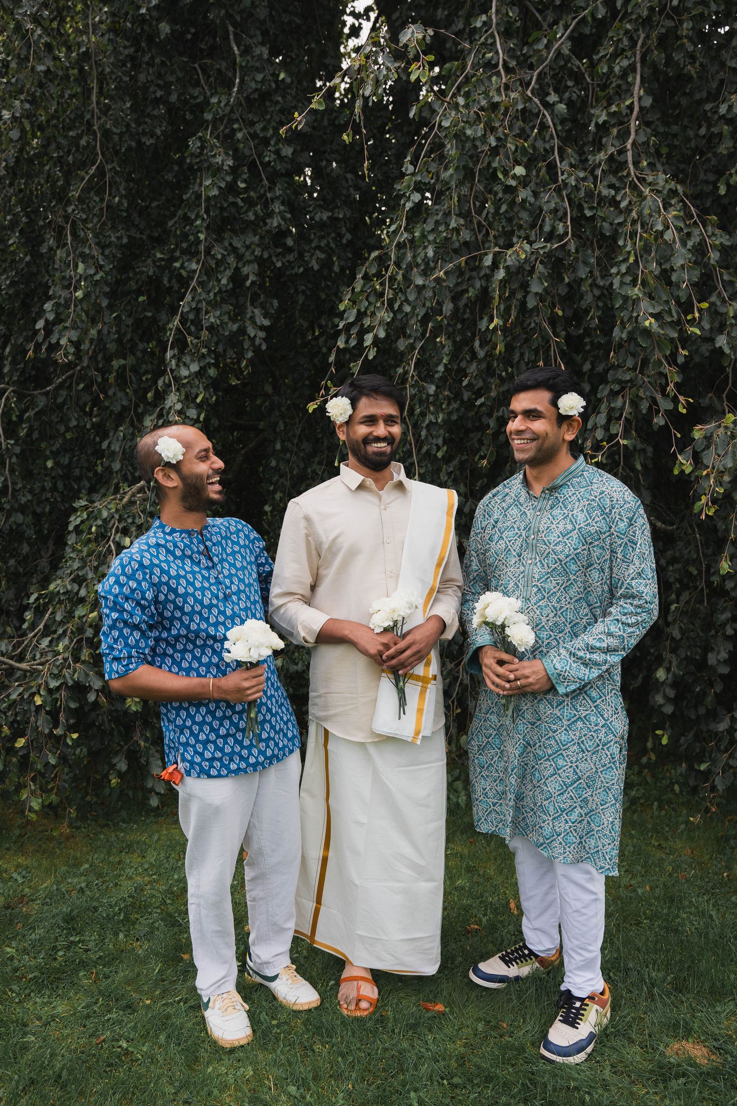 Three smiling men in kurtas and a traditional dhoti wear white flowers in their hair and hold bouquets under a weeping tree.