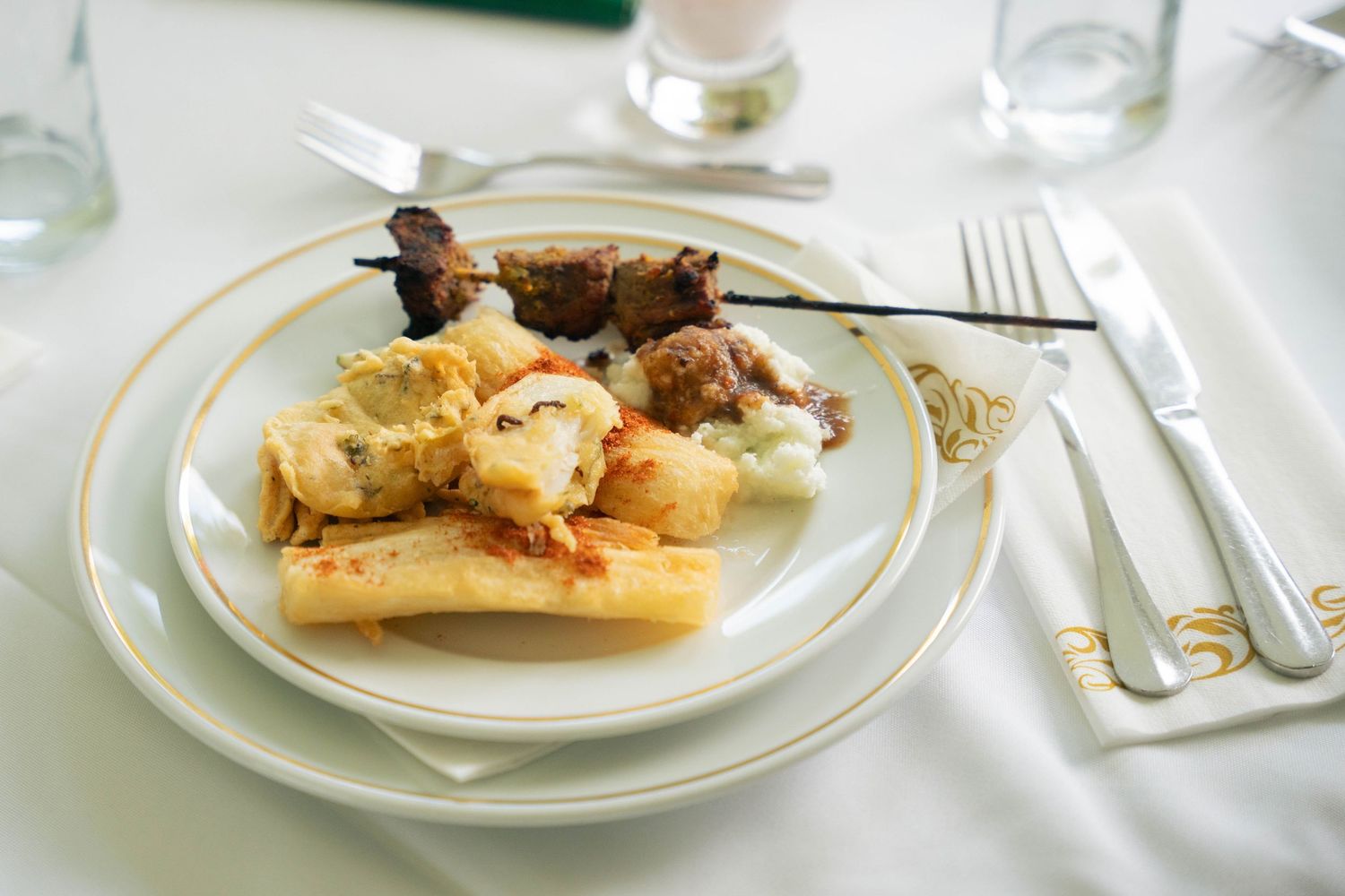 Plated meal with grilled meat skewer, stuffed dumplings and a creamy sauce on a gold-rimmed plate with silverware.