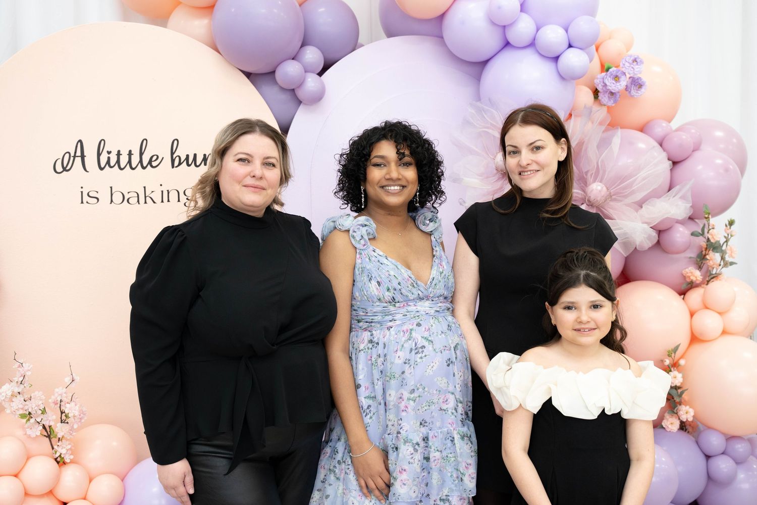 Four people pose in front of a pastel peach and lavender balloon display with a sign reading a little bun is baking at a baby shower.