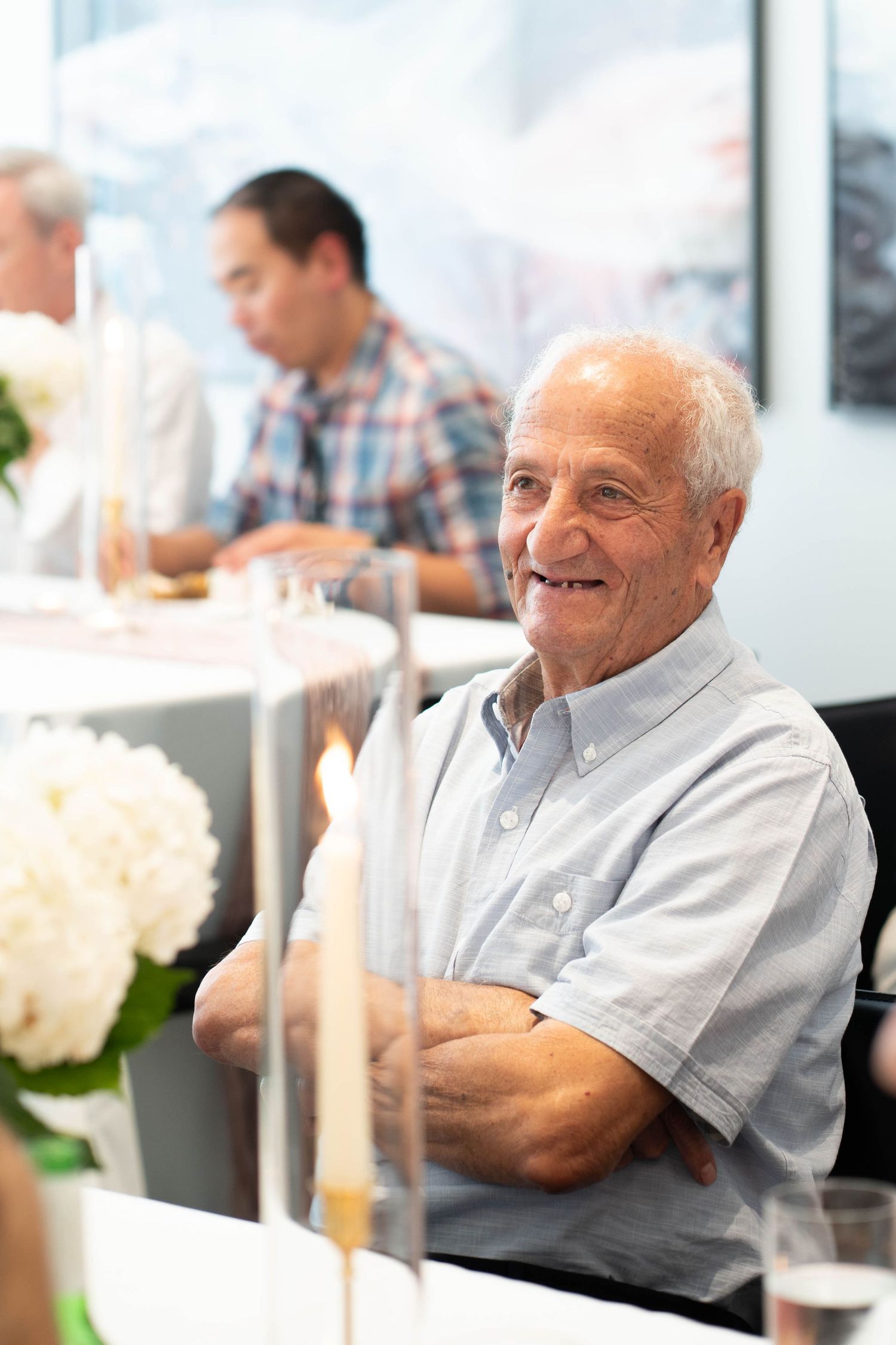 Smiling older man in a light blue button-up shirt sits with arms crossed at a table with candles and white flowers.
