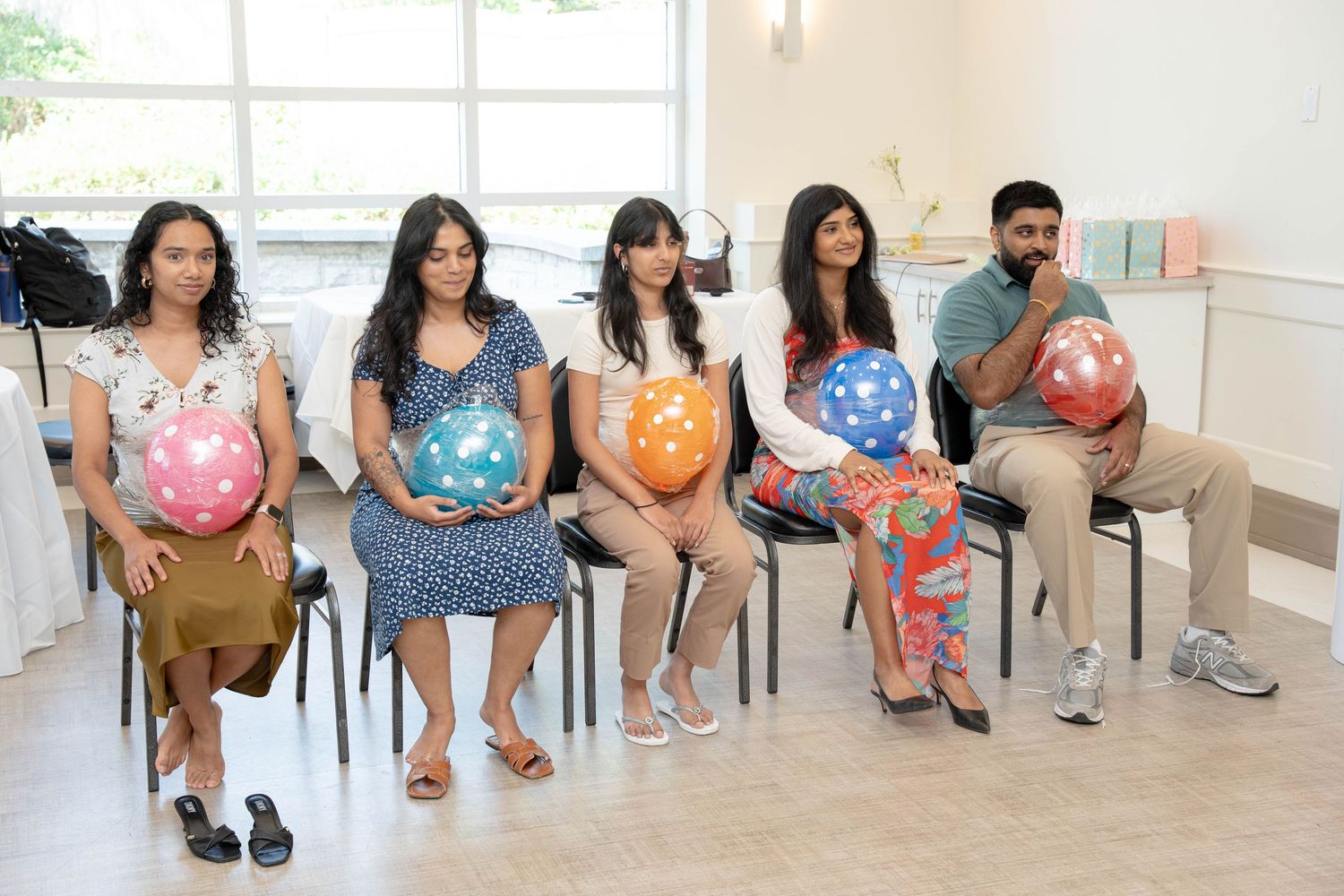 Five guests sit in a row holding colorful polka dot balloons while smiling at a baby shower celebration.