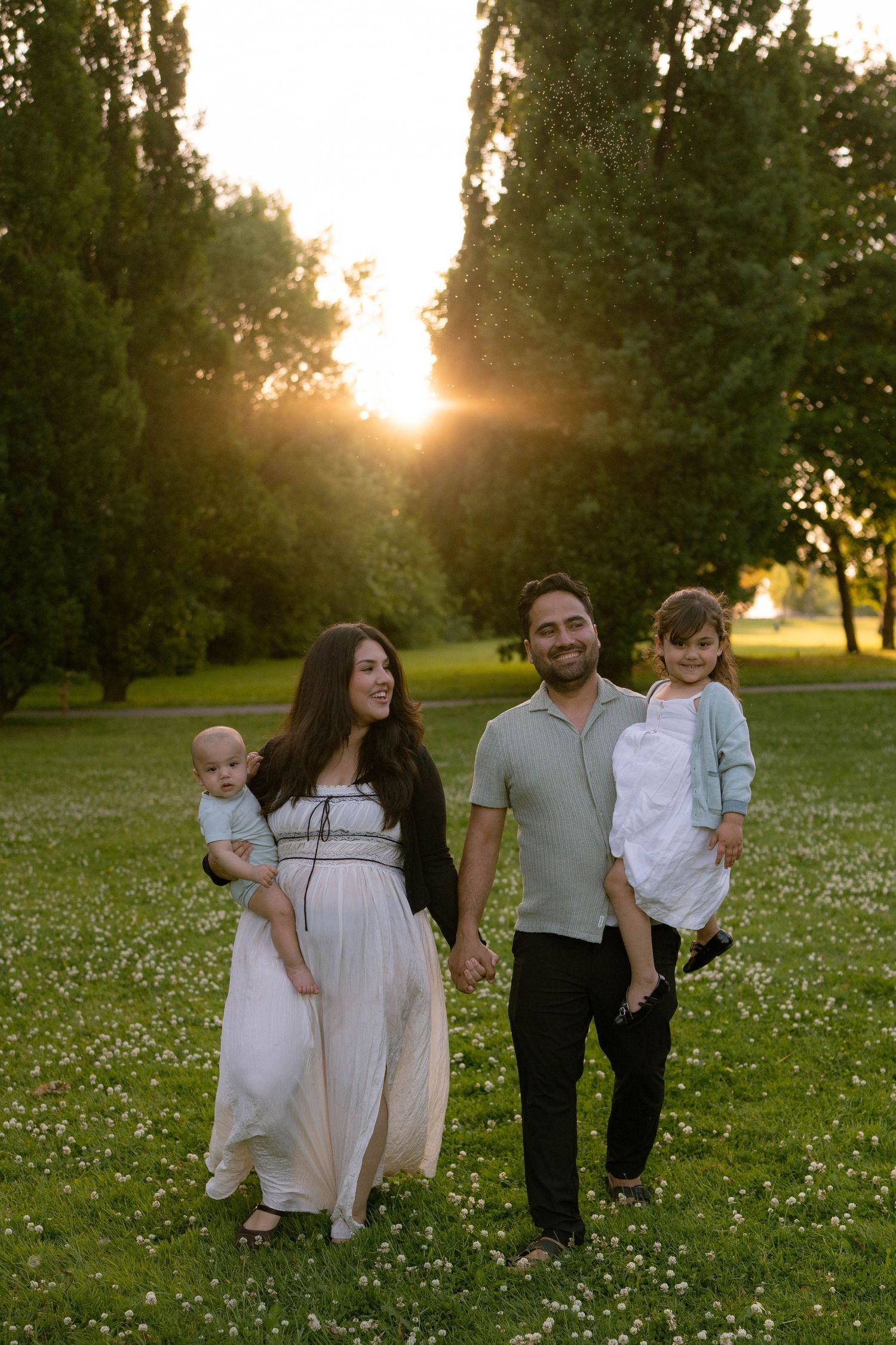 A family walks hand in hand through a flower-dotted meadow at sunset, with a baby and little girl in their arms.
