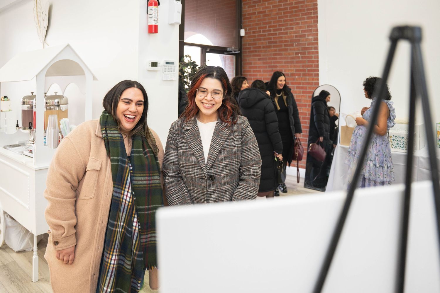 Two smiling women in coats pose near a white dessert cart as guests mingle at a baby shower event.