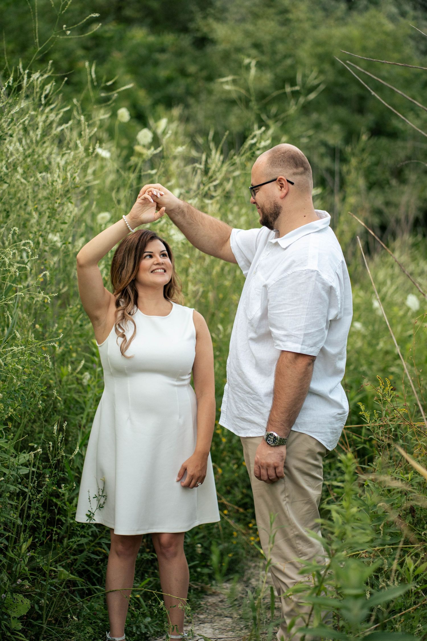 Woman in a white dress twirls under her partner's raised arm while dancing in a green summer field.