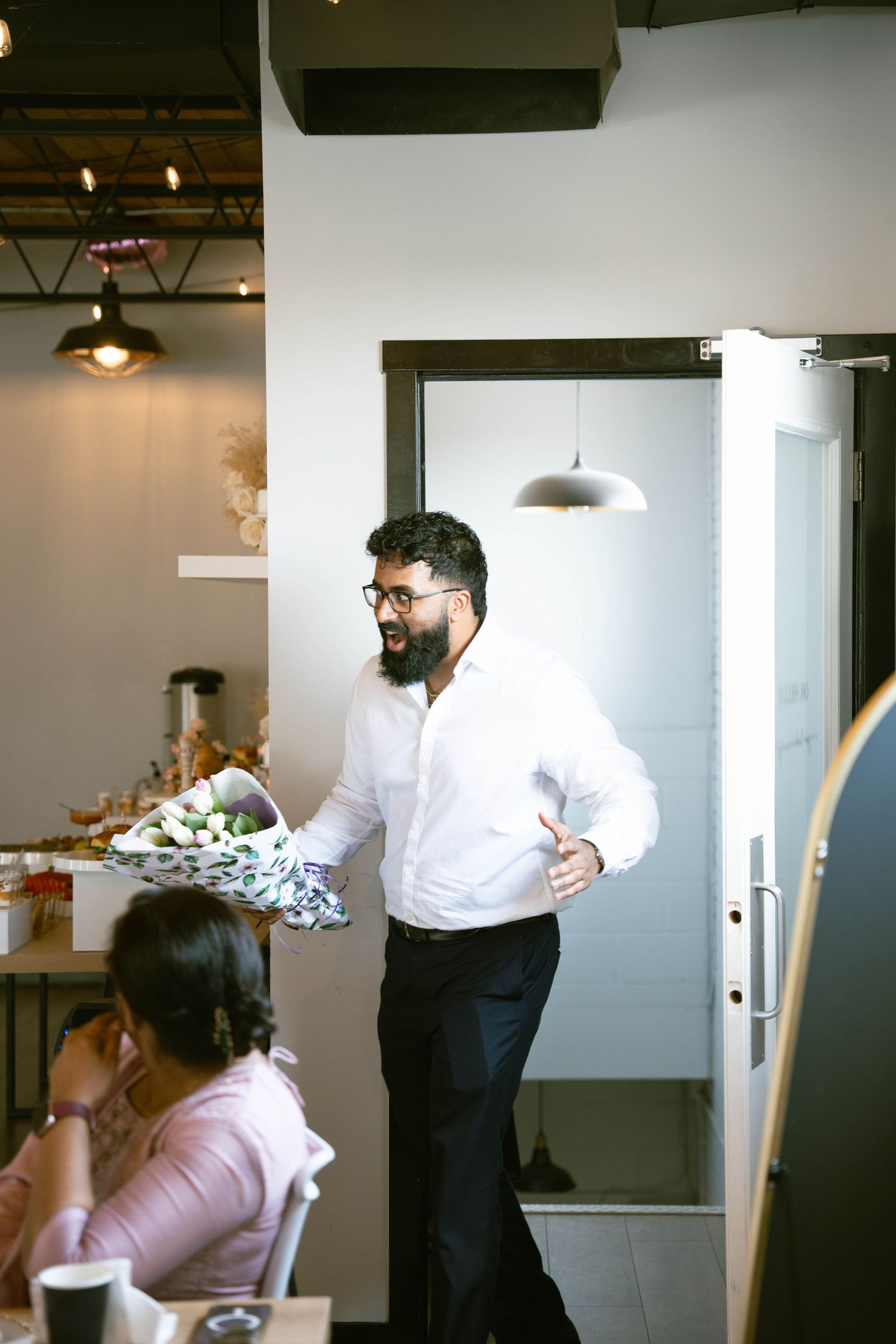 A bearded man in a white shirt enters a bridal shower holding a bouquet while a seated guest looks on.