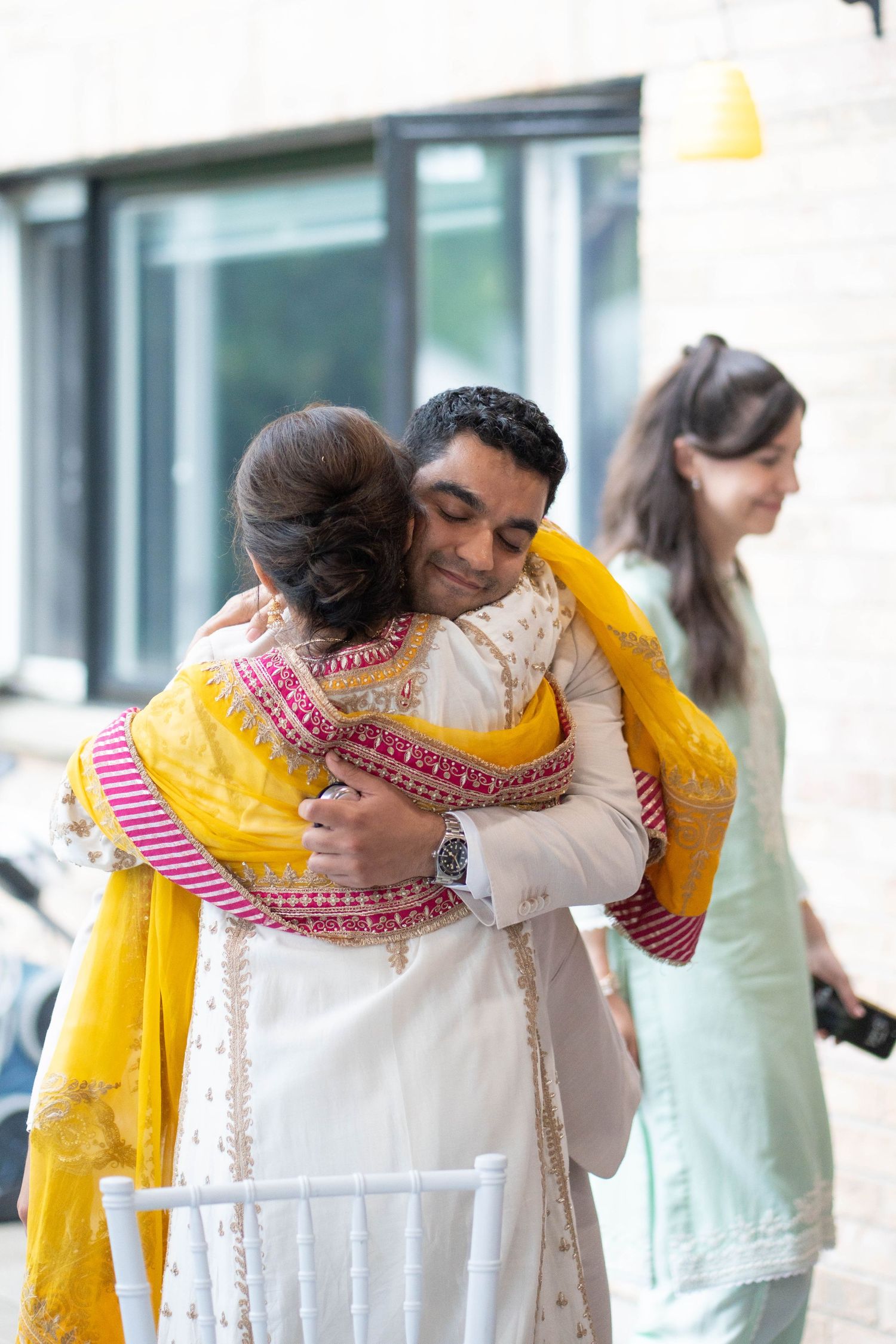 A man in a beige suit hugs a woman in a white and yellow embroidered outfit while a guest smiles in the background.