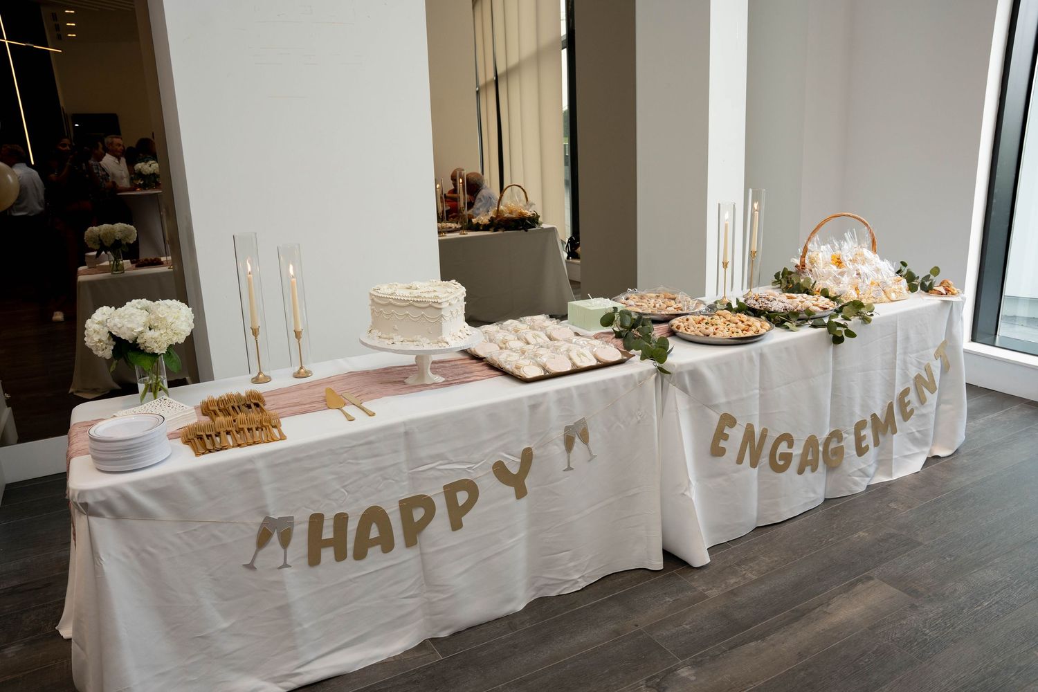 Dessert table with a white cake, treats, and floral garland beneath a banner that reads Happy Engagement.