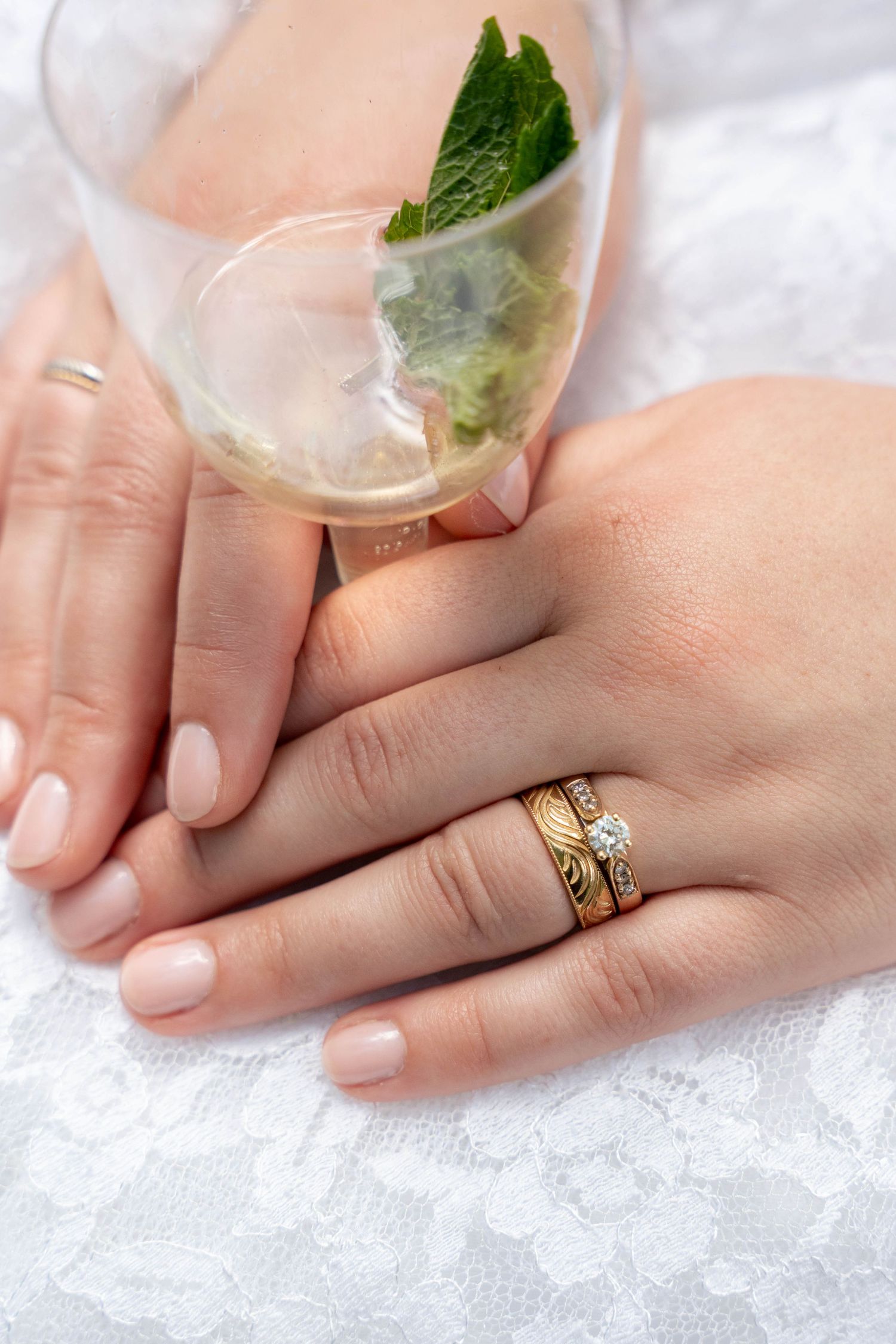 Close-up of a bride's hands wearing a gold wedding band and diamond ring, resting on lace beside a glass with a mint leaf.