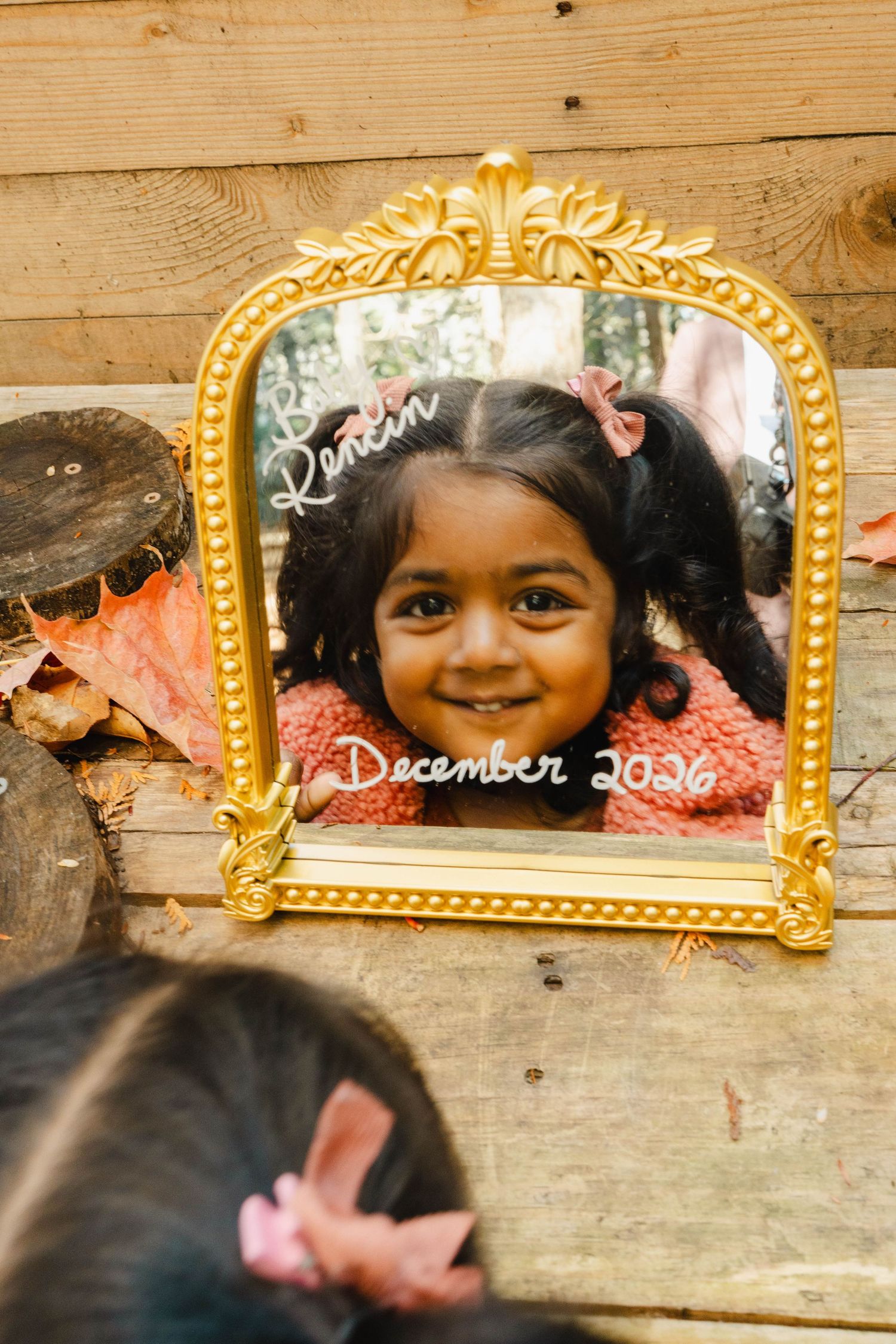 Smiling toddler girl with pink hair bows is reflected in a gold mirror marked with a baby name and December 2026 due date.