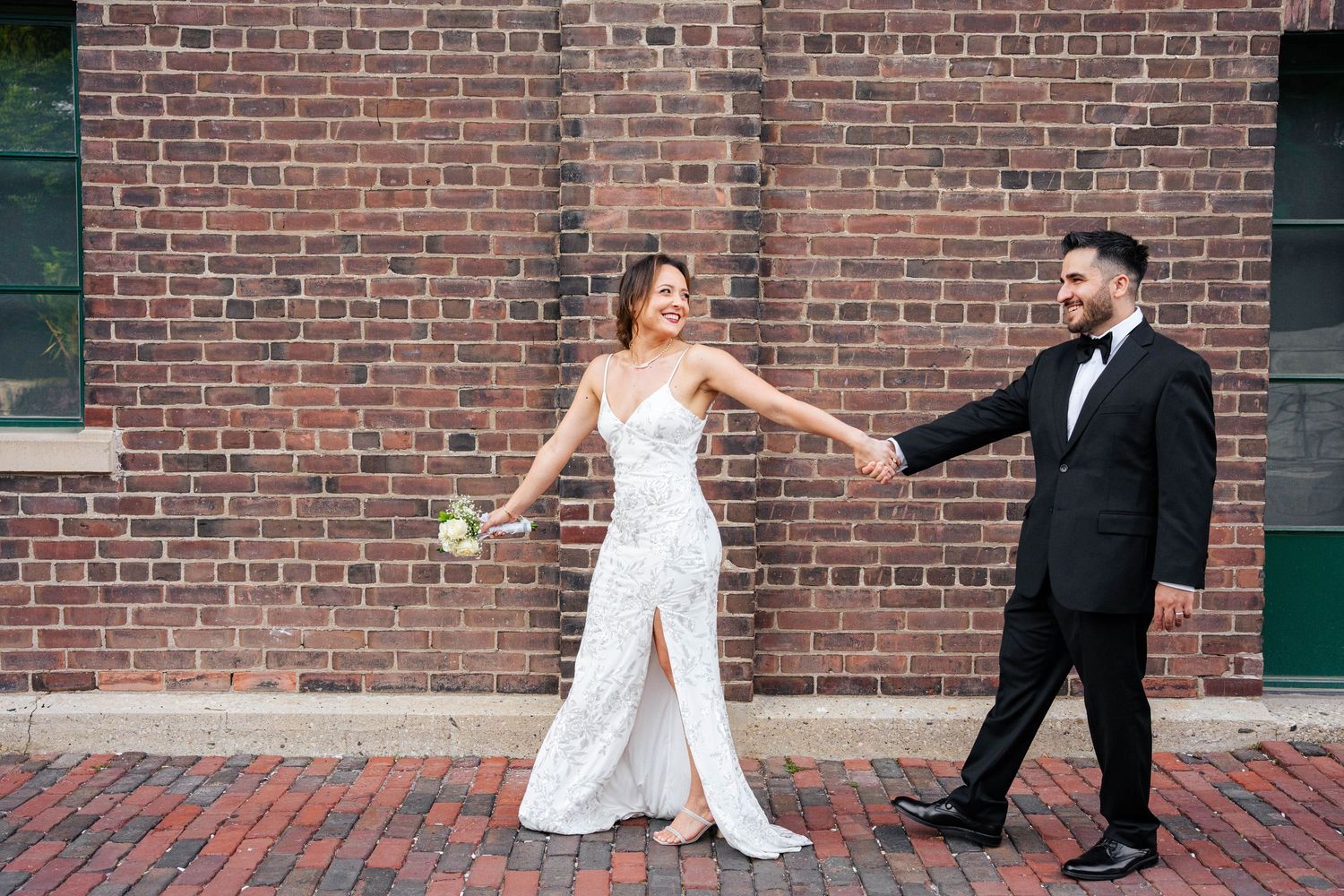 Bride in a white lace gown reaches for her groom's hand as they stand on a brick sidewalk beside a historic red brick building.