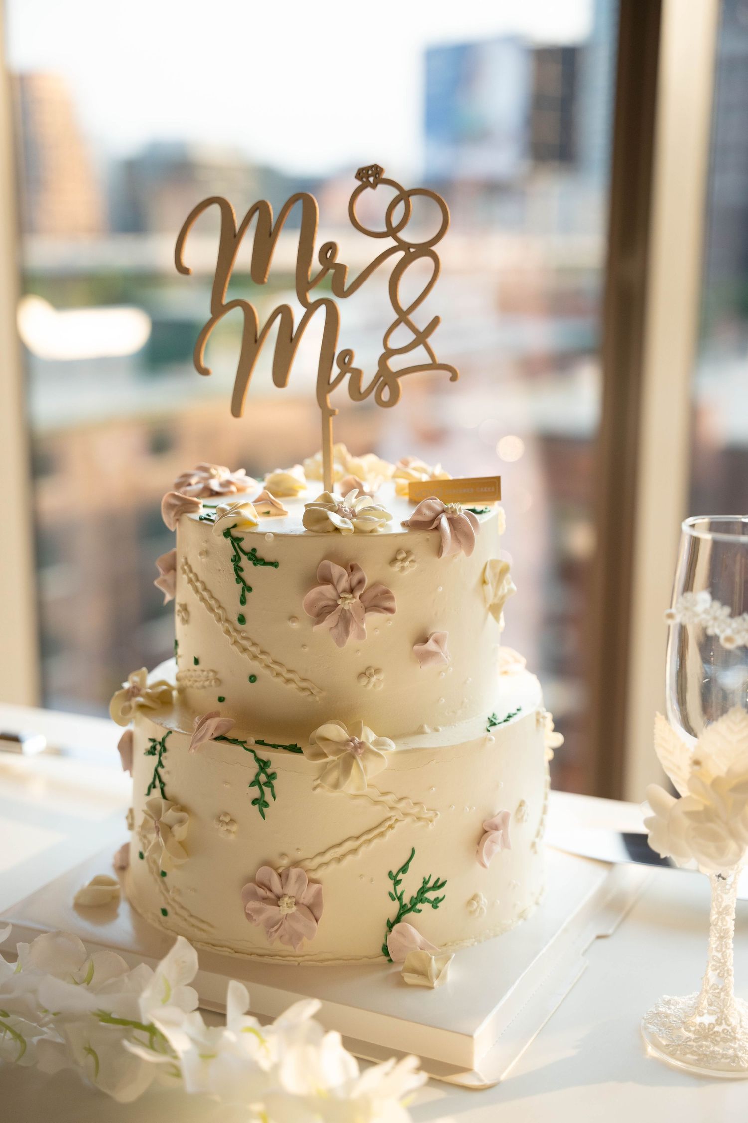 Two-tier white wedding cake with pink sugar flowers and a gold Mr and Mrs topper beside a champagne flute and city skyline.
