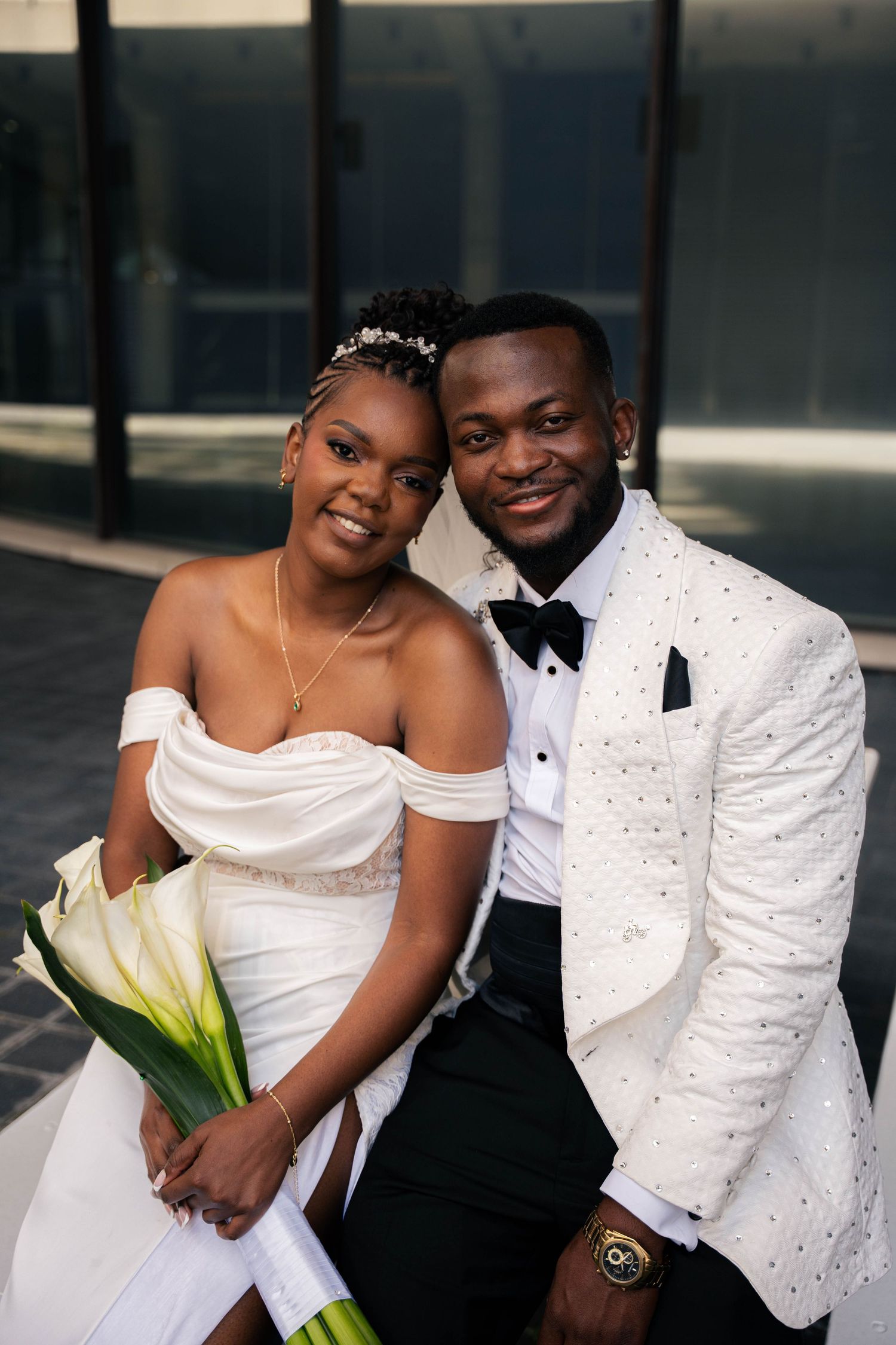 Smiling bride and groom sit close together, she holding white calla lilies, he wearing an ivory jacket with a black bow tie.