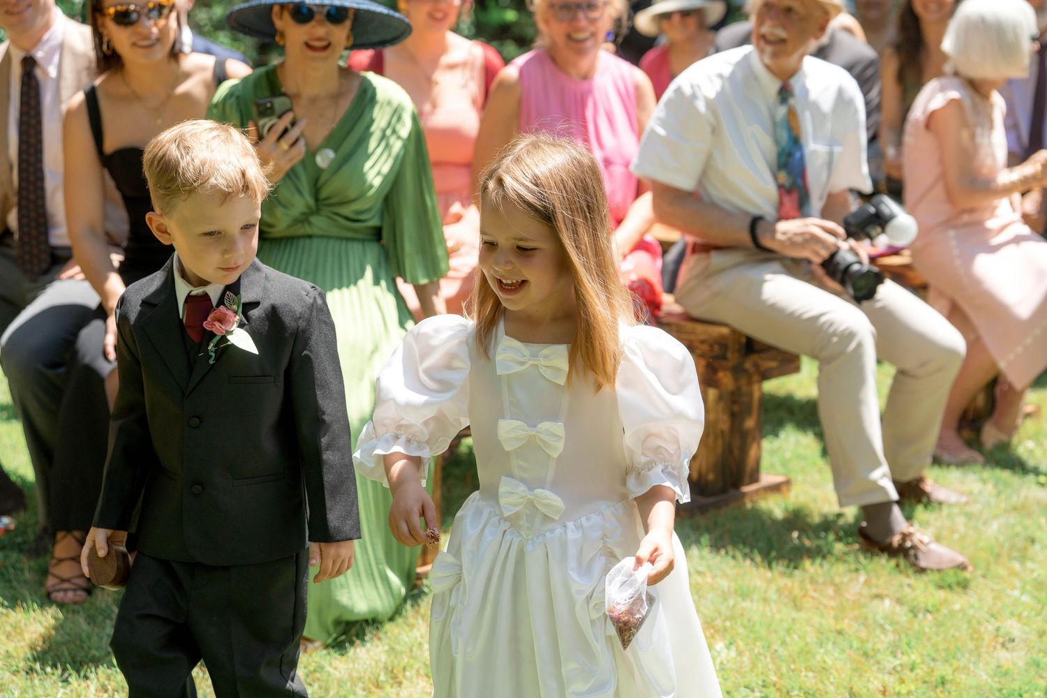 A young ring bearer in a dark suit and a flower girl in a white dress walk together at an outdoor wedding ceremony.