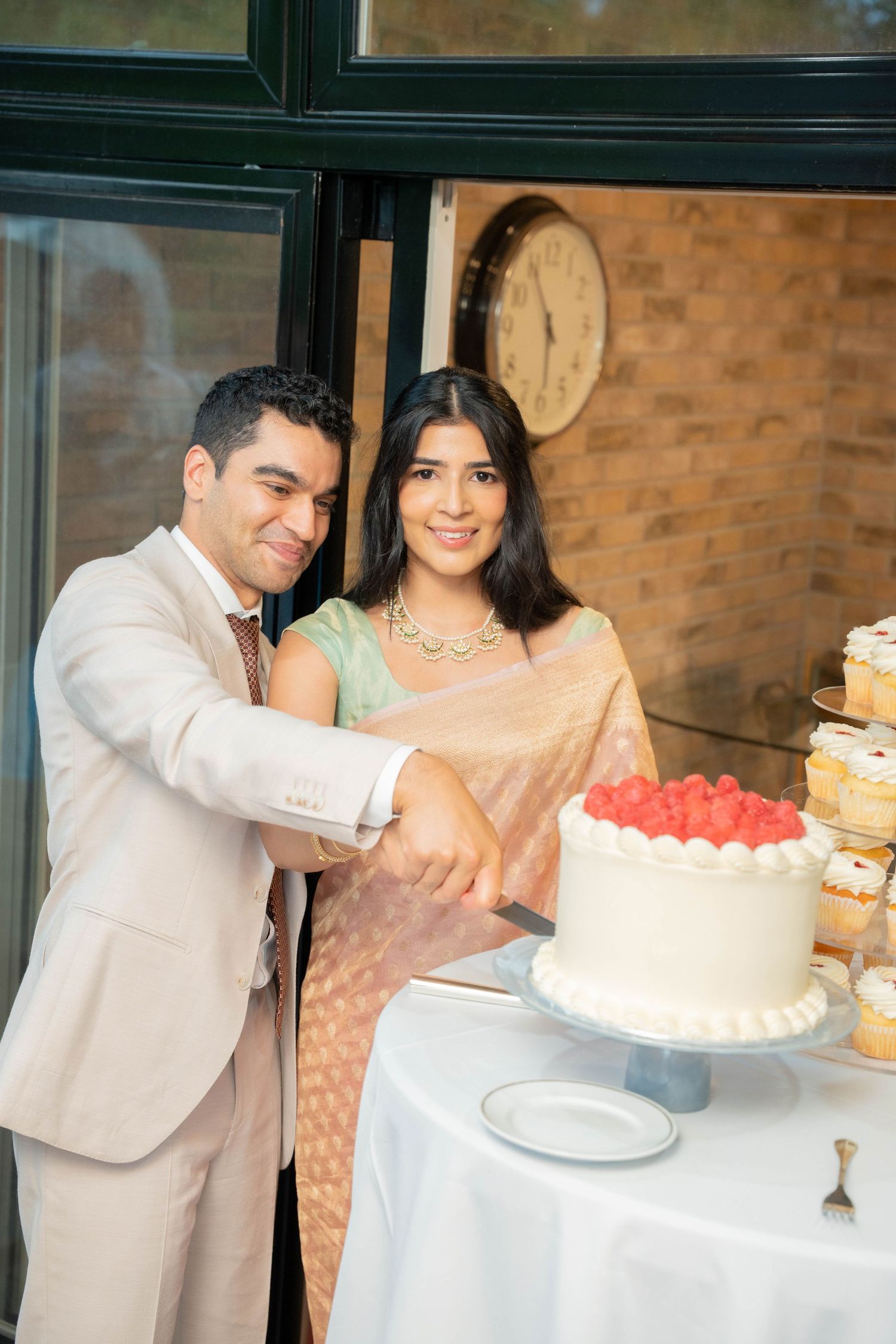 A smiling couple in a beige suit and peach saree cut a white cake topped with fresh strawberries beside a dessert display.