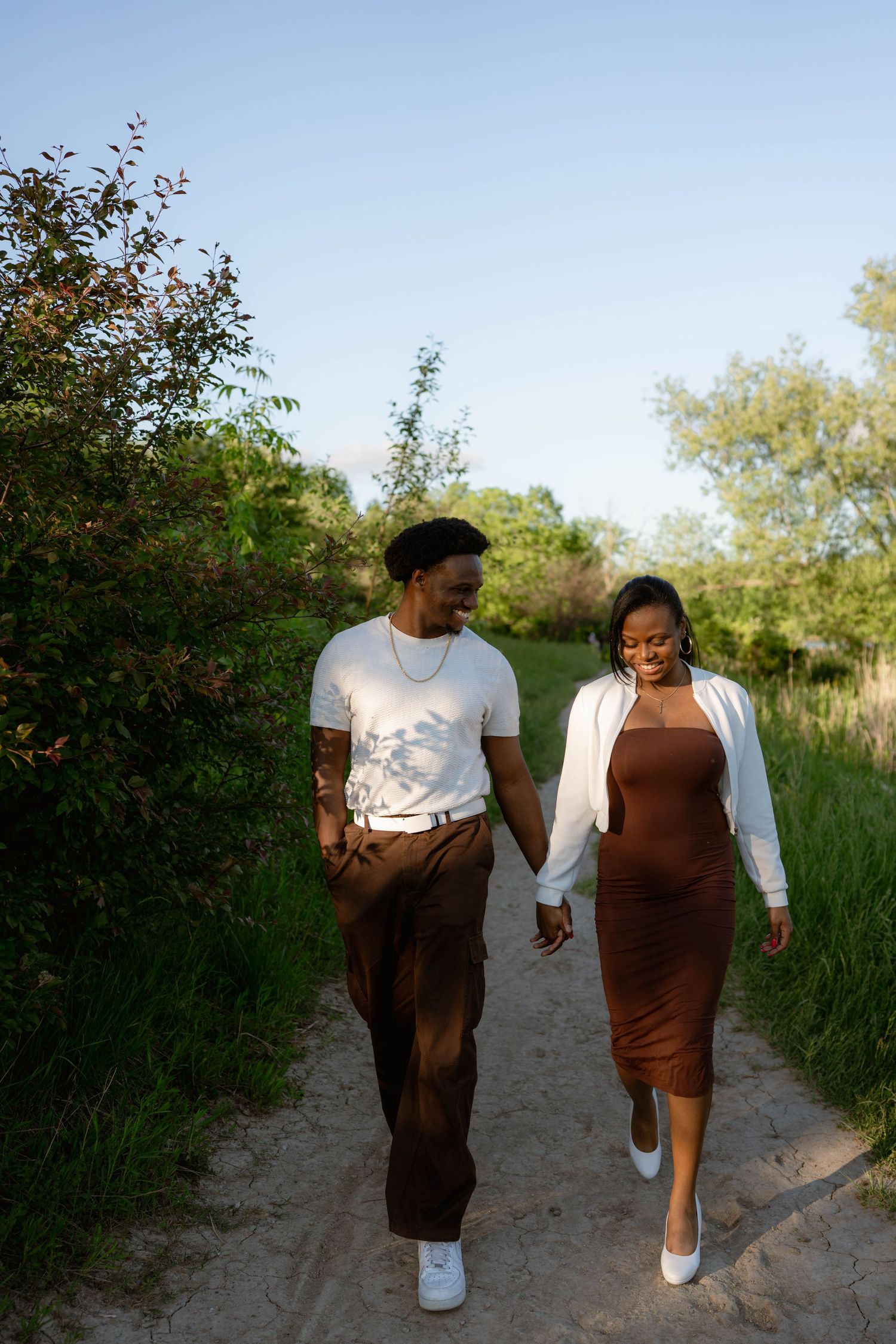 An engaged couple holds hands and smiles while walking down a sandy nature path surrounded by greenery.
