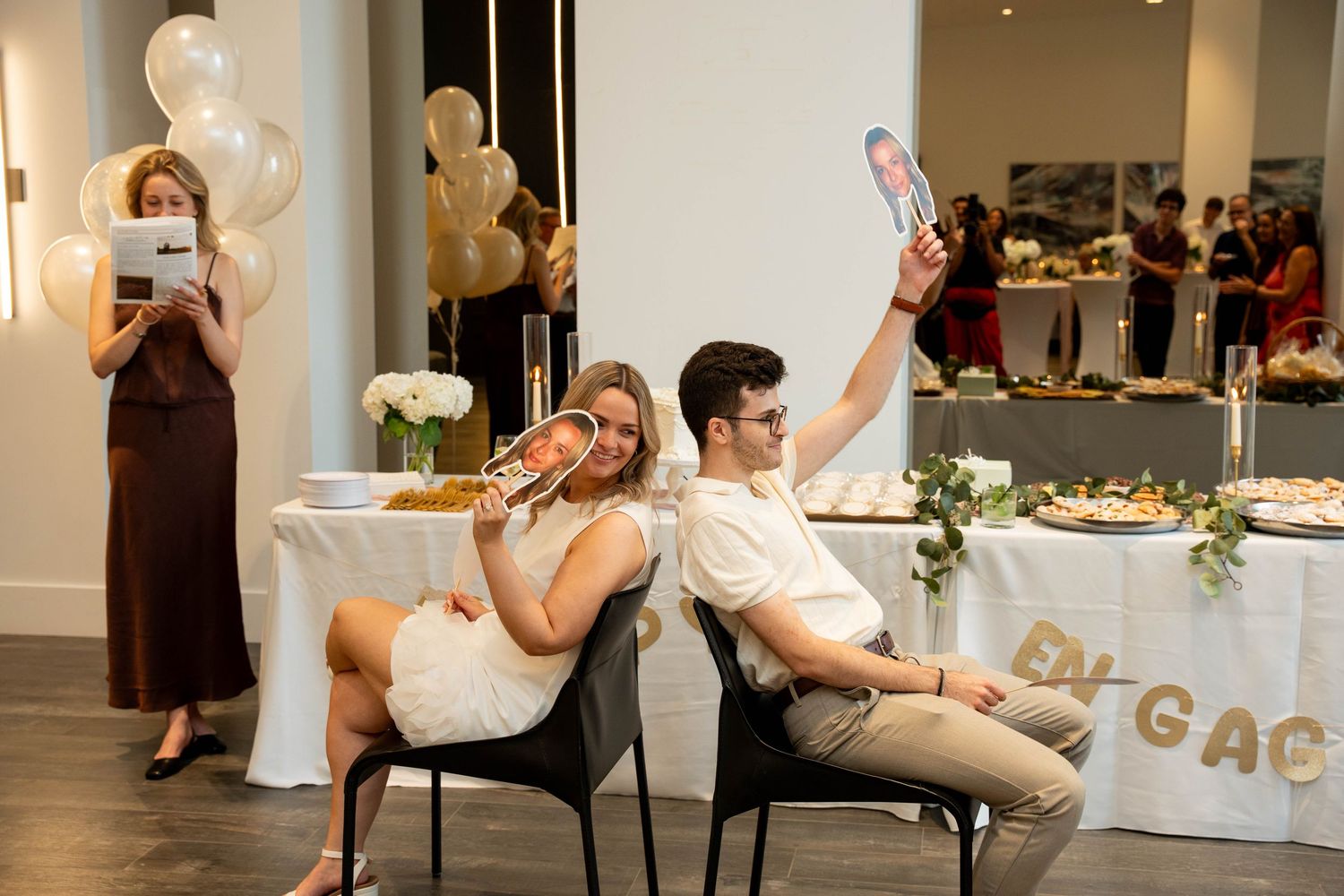 Guests at an engagement party hold up cardboard face cutouts on sticks while a woman reads a card near gold balloons and a buffet.