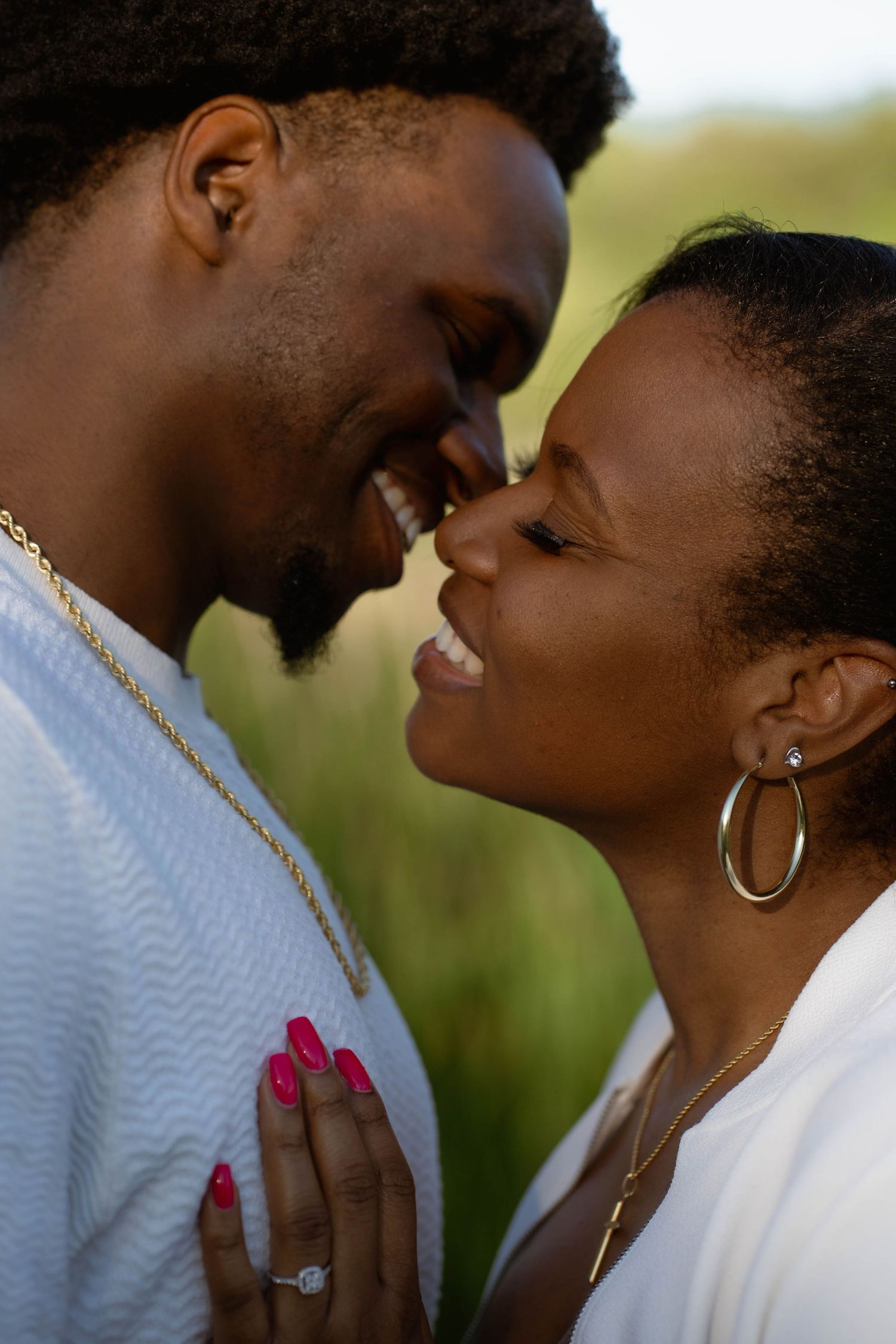 A joyful couple nuzzles noses and smiles during an outdoor engagement photoshoot, her ring visible on his chest.