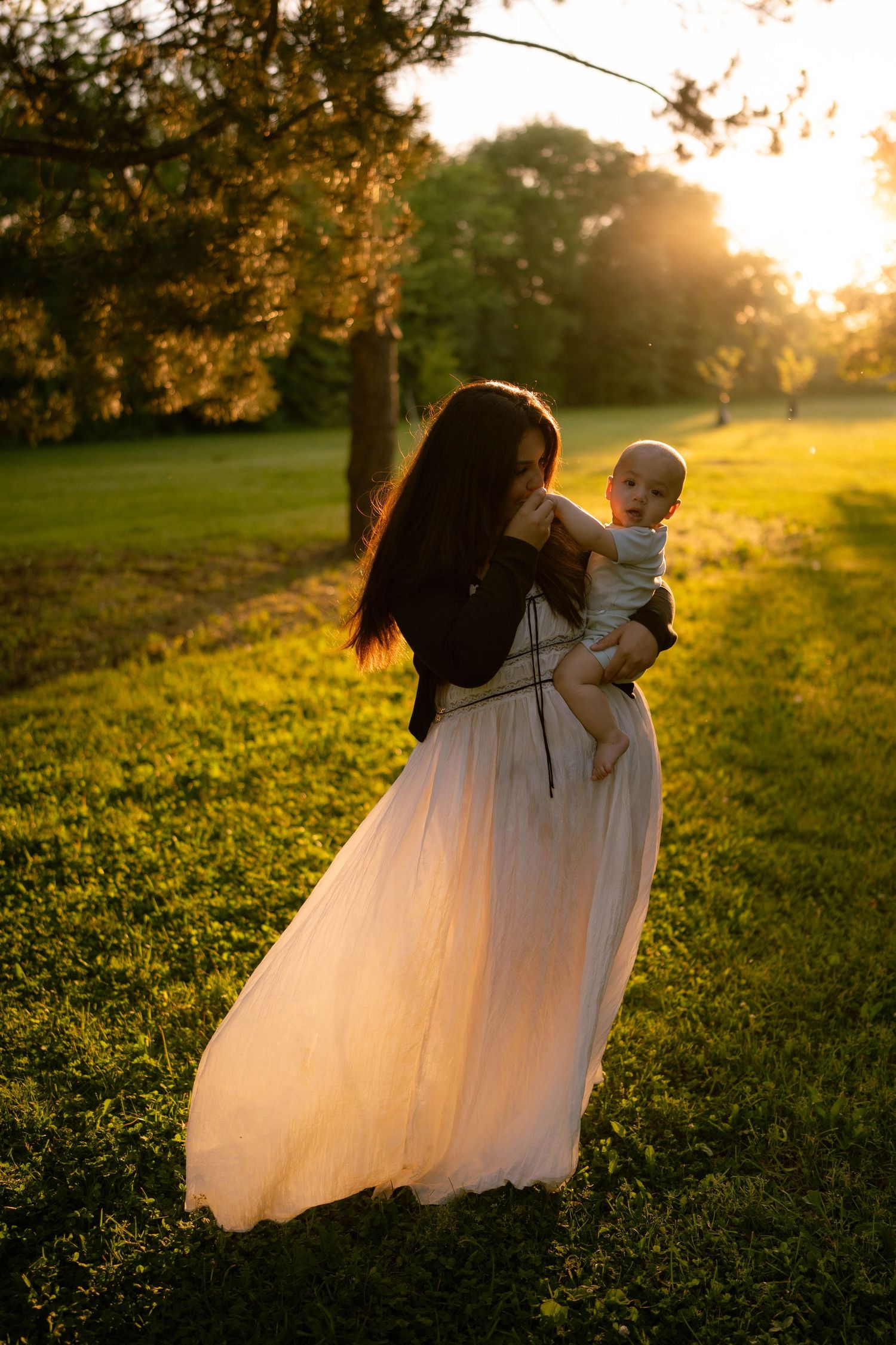 A mother with long dark hair holds her baby while walking through a golden sunset-lit park in a flowing white dress.