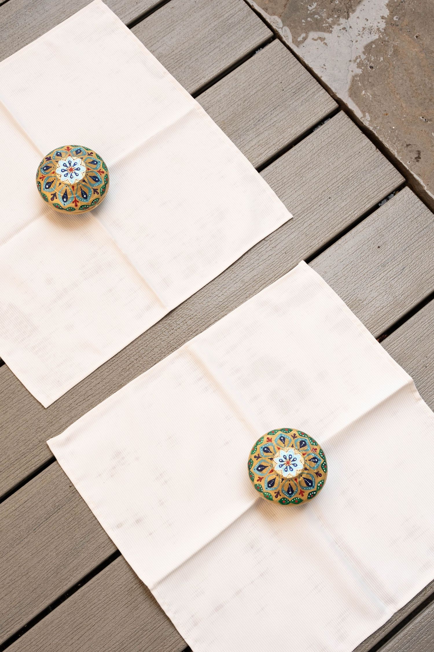 Two cream linen napkins on a wooden deck, each topped with an ornate hand-painted decorative ceramic knob.