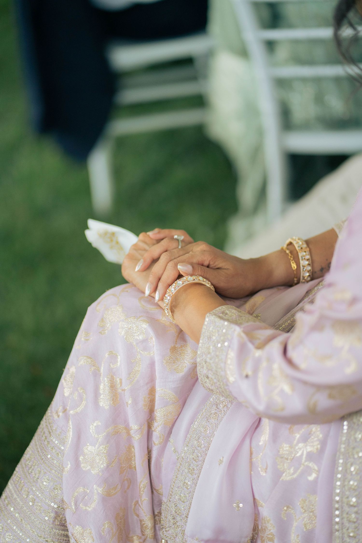 Close-up of a woman's hands with gold bangles resting on a pale lavender embroidered outfit at an outdoor wedding.